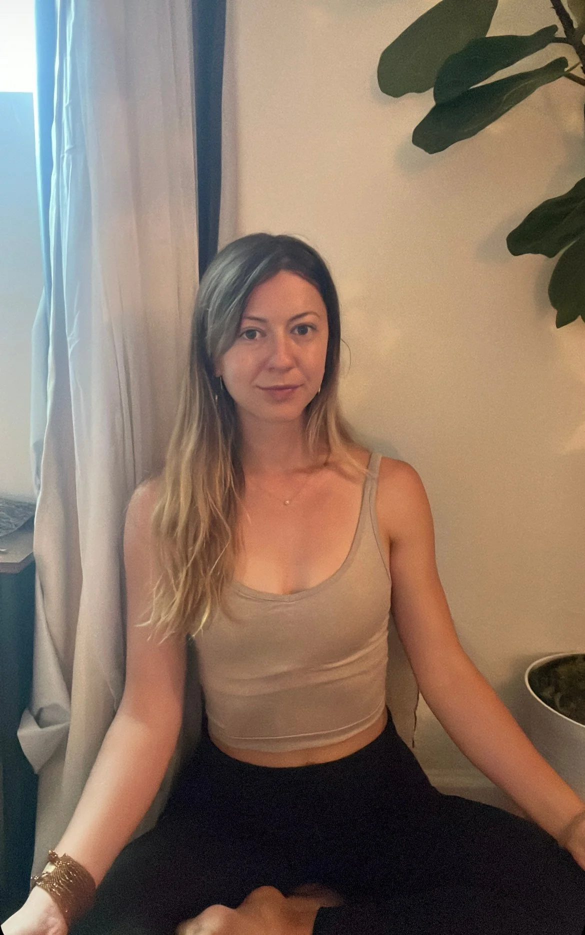 Woman sitting in meditation pose next to potted plant indoors.