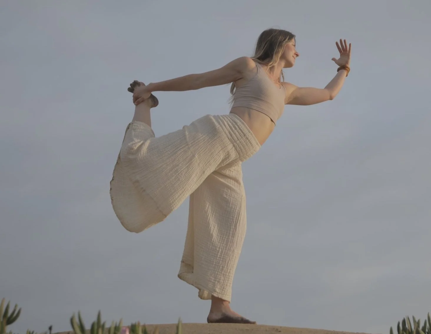 Woman in yoga pose outdoors, wearing a beige top and wide-legged pants, balancing on one leg with the sky in the background.