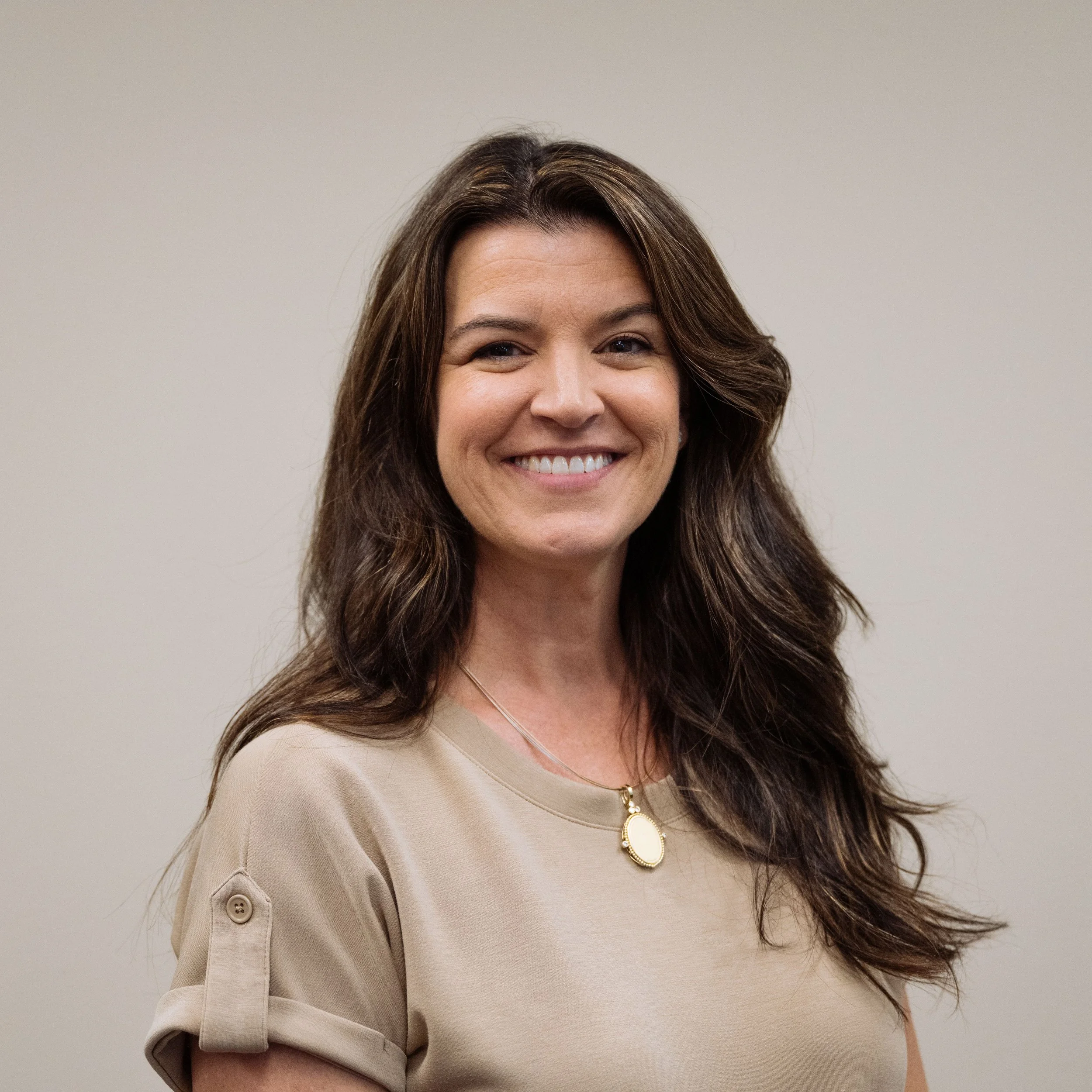 Smiling woman with long brown hair wearing a beige shirt and pendant necklace against a plain background.