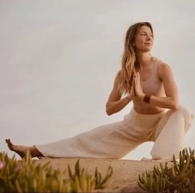 A woman practicing yoga outdoors on a sandy surface, sitting in a side stretch pose with hands pressed together in prayer position.