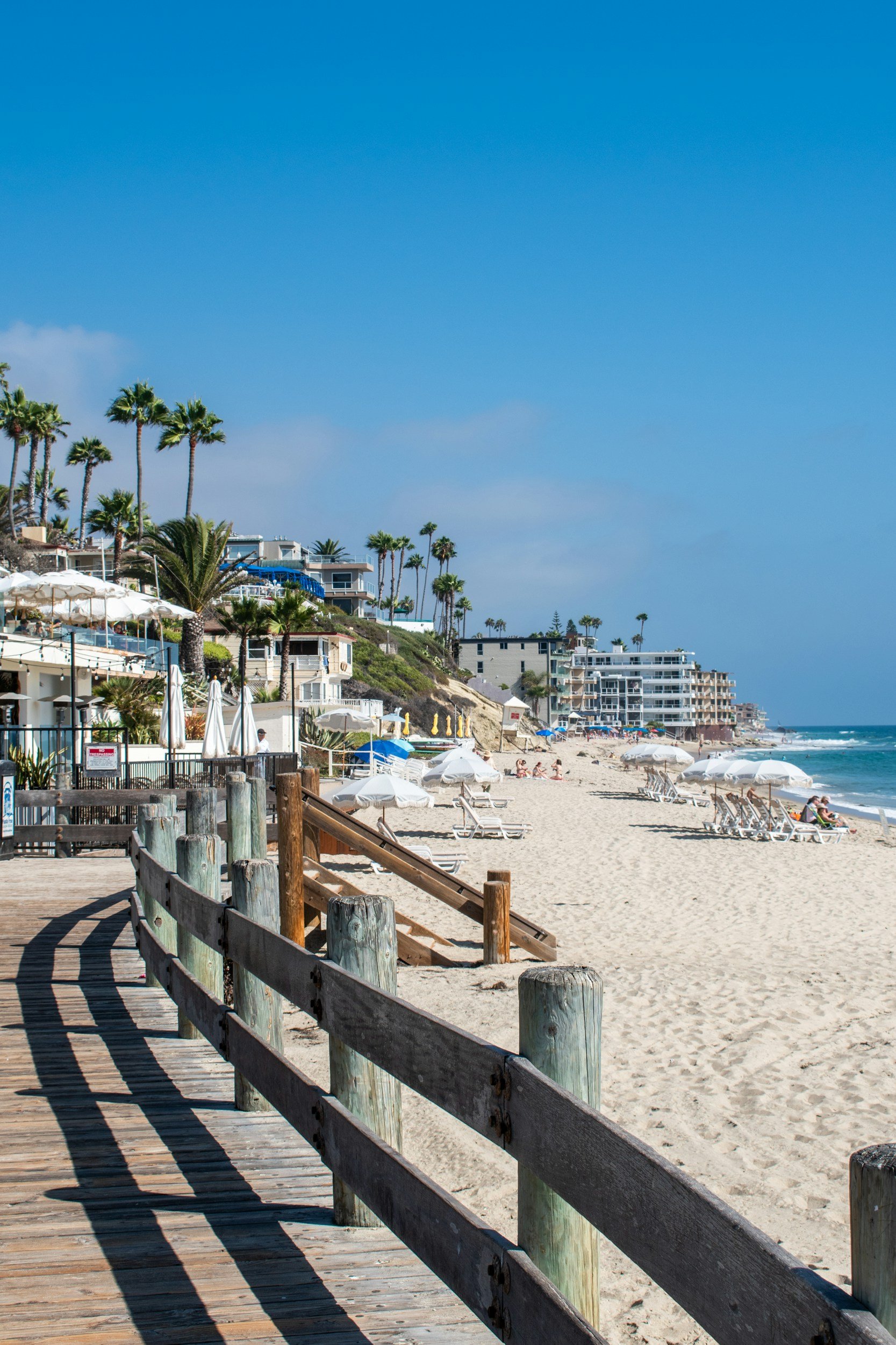 Beach scene with wooden boardwalk and railing leading to sandy shore, beach chairs, umbrellas, luxury beachfront homes, palm trees, and blue sky with a few clouds.