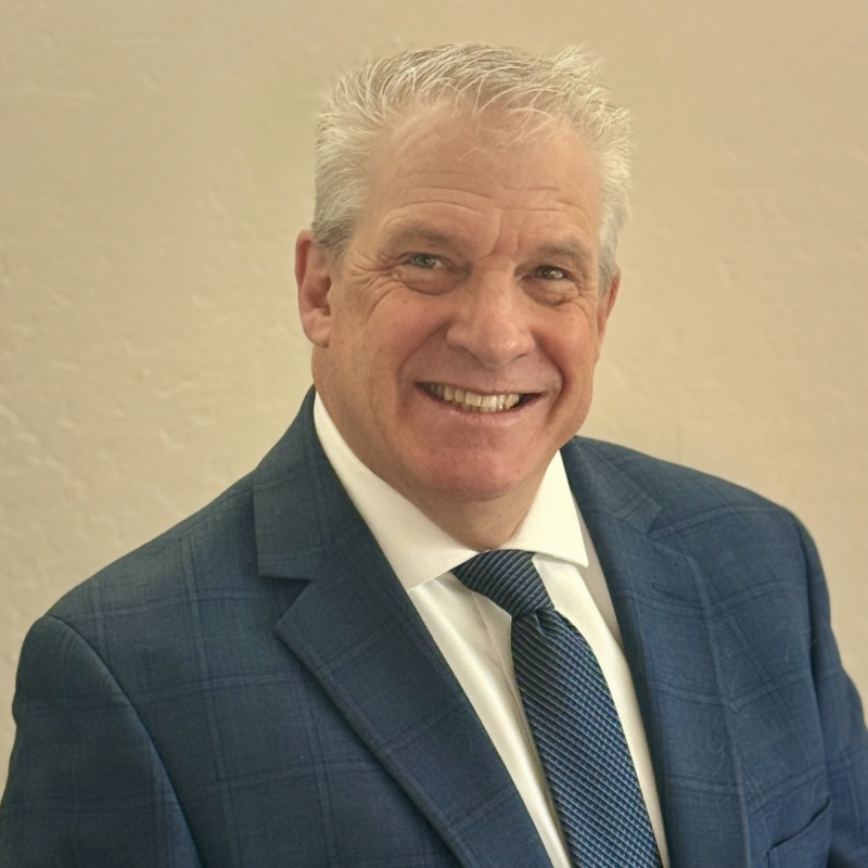 Smiling middle-aged man with gray hair in a dark blue suit, white shirt, and striped tie, standing against a plain light-colored wall.