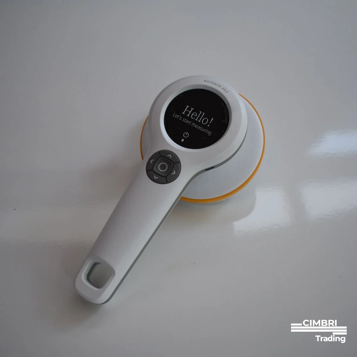 A medical thermometer with a round digital display showing the message 'Hello! Let's start measuring.' The thermometer is white with black buttons and is placed on a yellow base on a white surface.