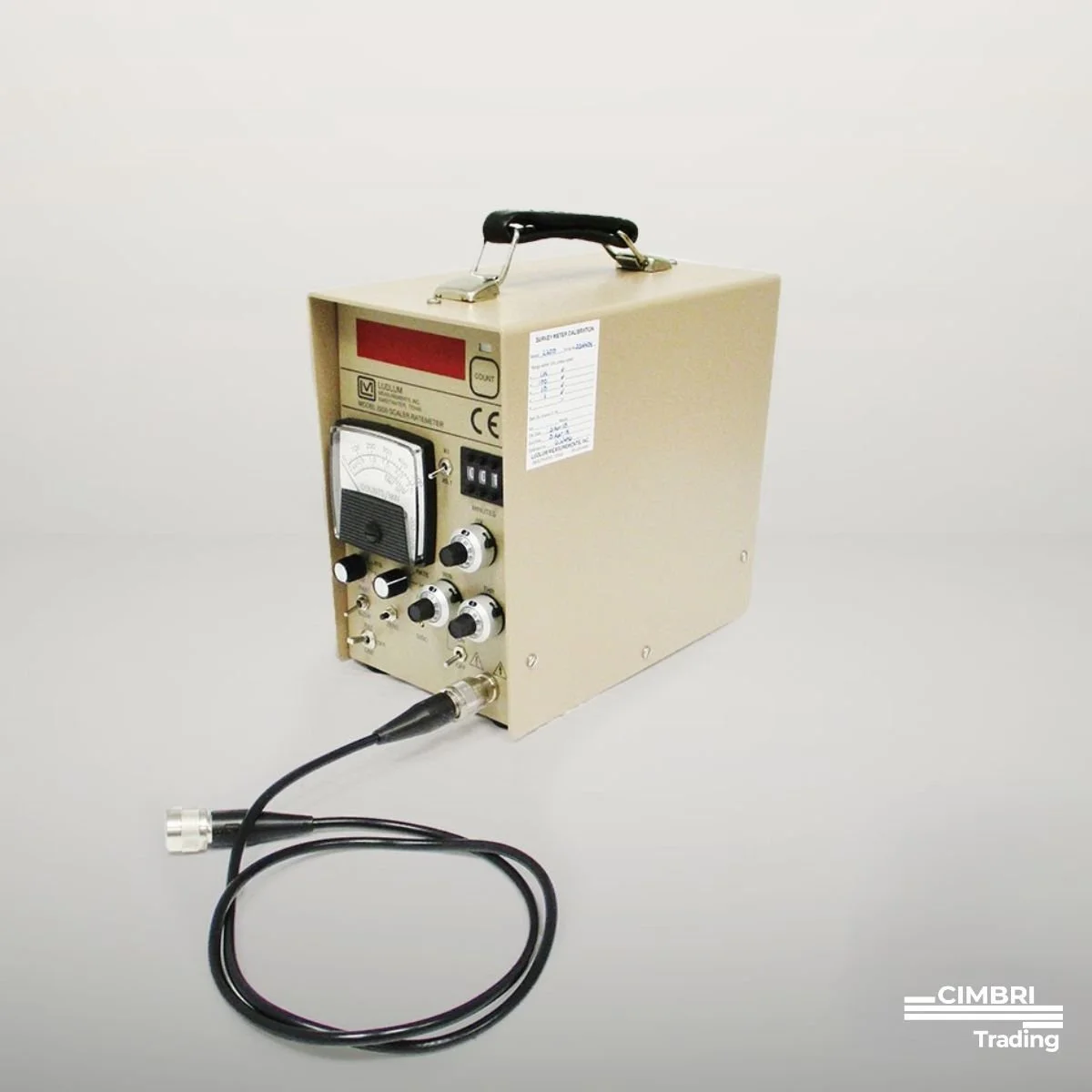 A vintage electronic device with a handle, knobs, a dial, an analog gauge, and a connected cable on a plain background.