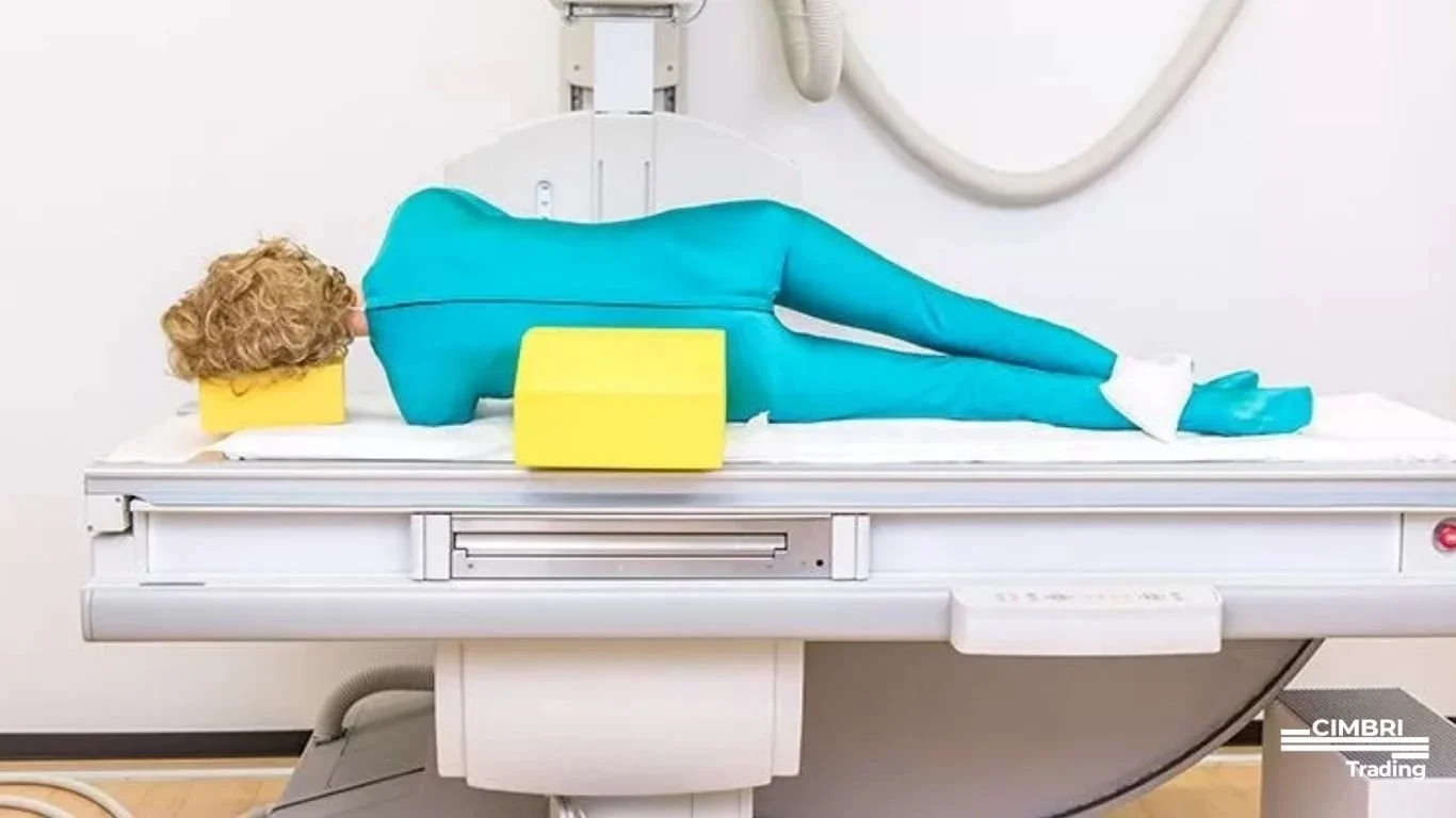A person lying on a medical imaging table, wearing protective blue clothing, with yellow cushioning support.