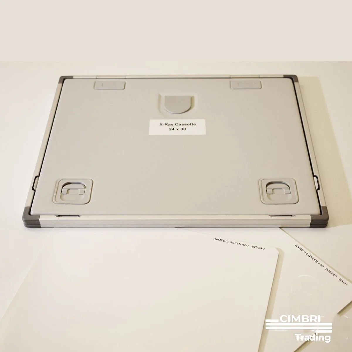 Flat white X-ray cassette box labeled "X-Ray Cassette 24 x 30" on a white surface with several white paper sheets underneath, some marked "HAMEDIC GREEN 400."