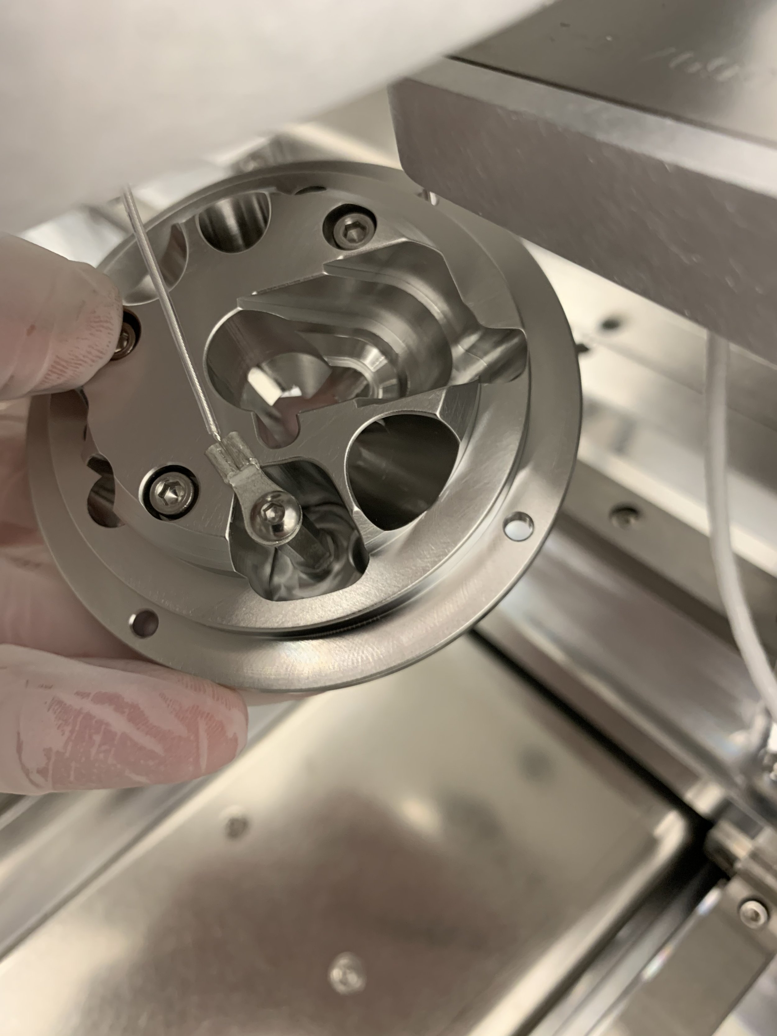 Close-up of a metal mechanical part with screws, being handled with a gloved hand, possibly a component of a machine or equipment, inside an industrial setting.