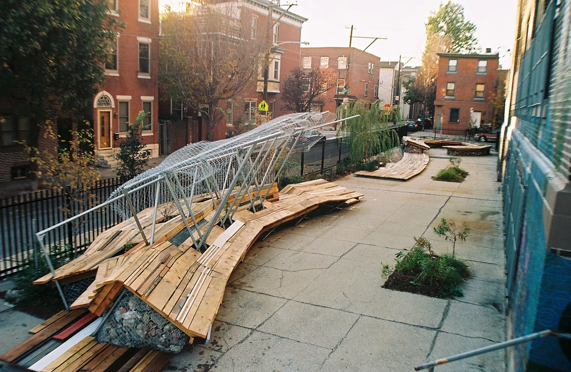 Urban sidewalk with wooden benches and metal railings, construction scaffolding leaning on the benches, small planted trees along the sidewalk, brick apartment buildings in the background, and a construction barrier on the right side.