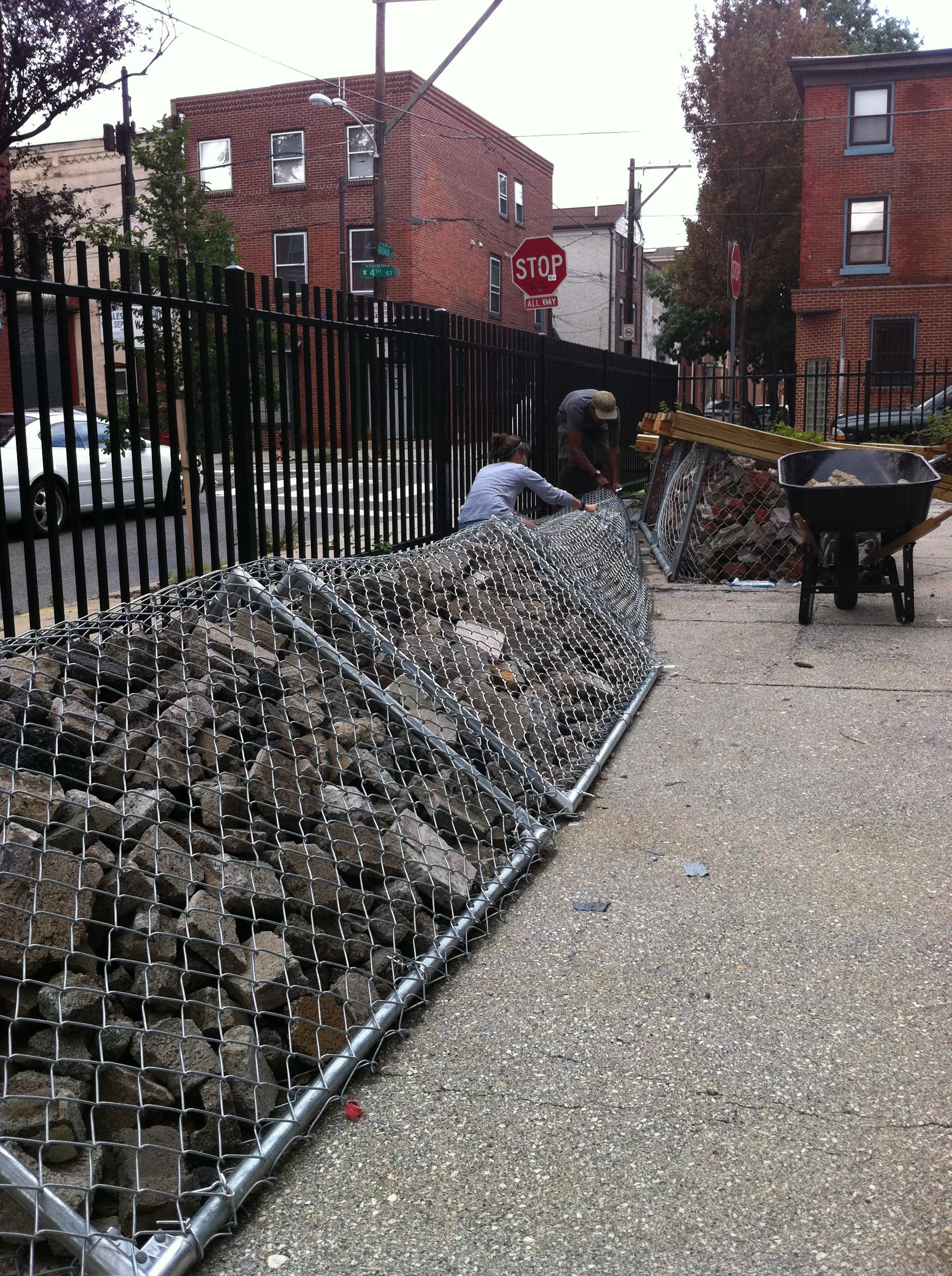 Two people working with wire fencing and rocks on a city sidewalk near a black metal fence. One person is kneeling and the other is bending over, with a wheelbarrow holding a mix of rocks and construction materials nearby. Behind them, there are bric