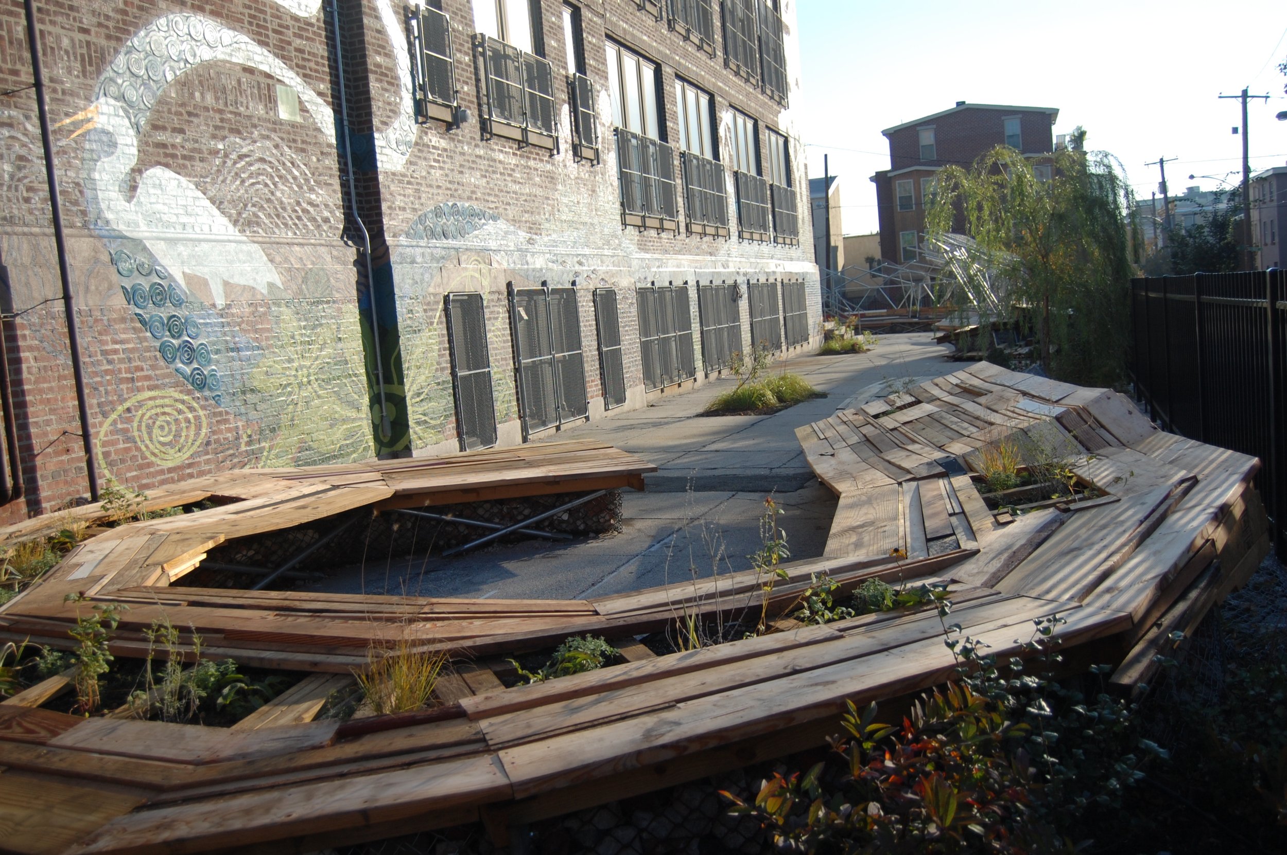 An outdoor urban space with a stepped wooden pathway surrounded by plants, next to a brick building with a mural of a bird painted on the wall.