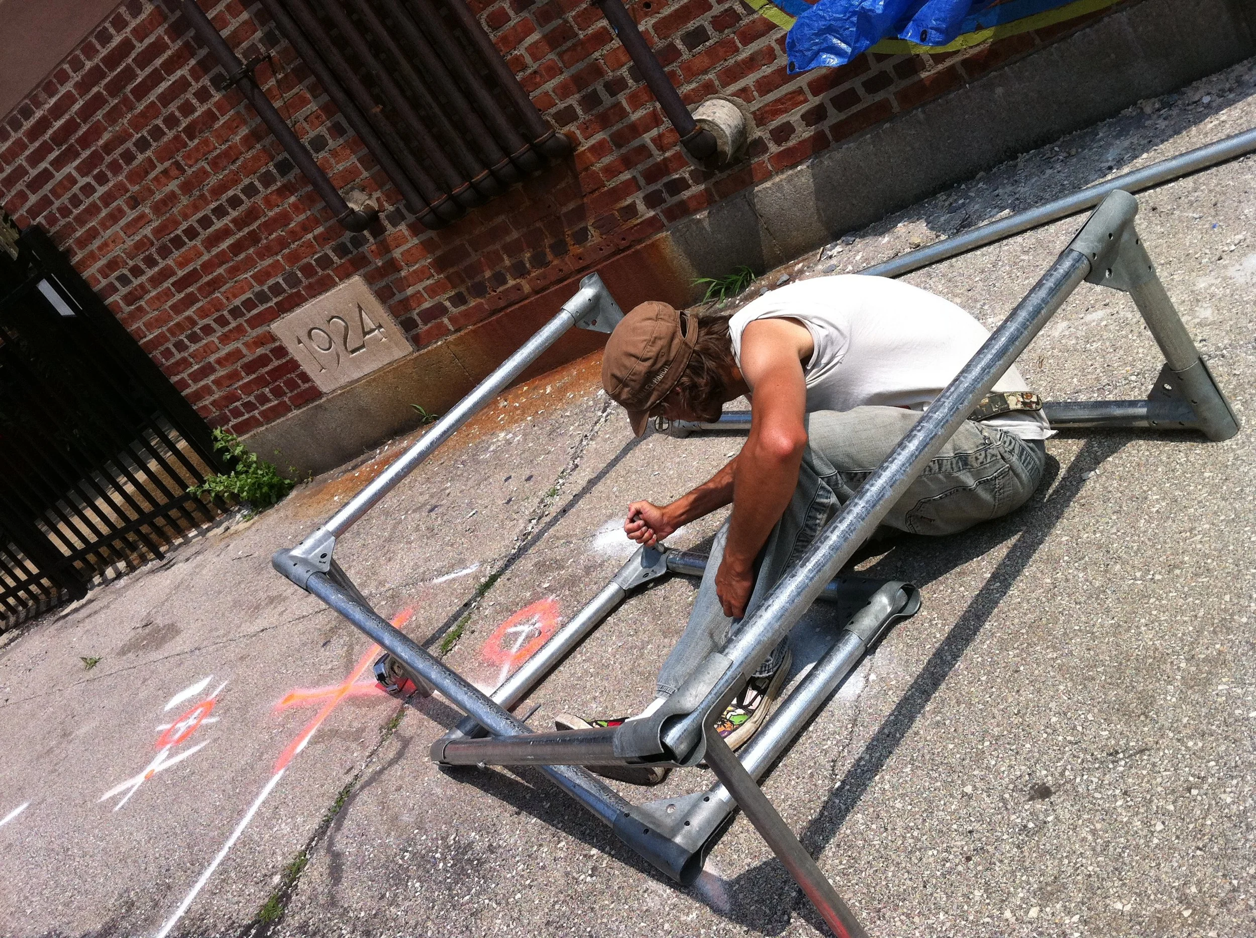 A man with long hair, wearing a brown cap, white t-shirt, and gray pants, kneeling on a sidewalk assembling a metal frame next to a brick wall with the number 1924 and painted markings on the ground.