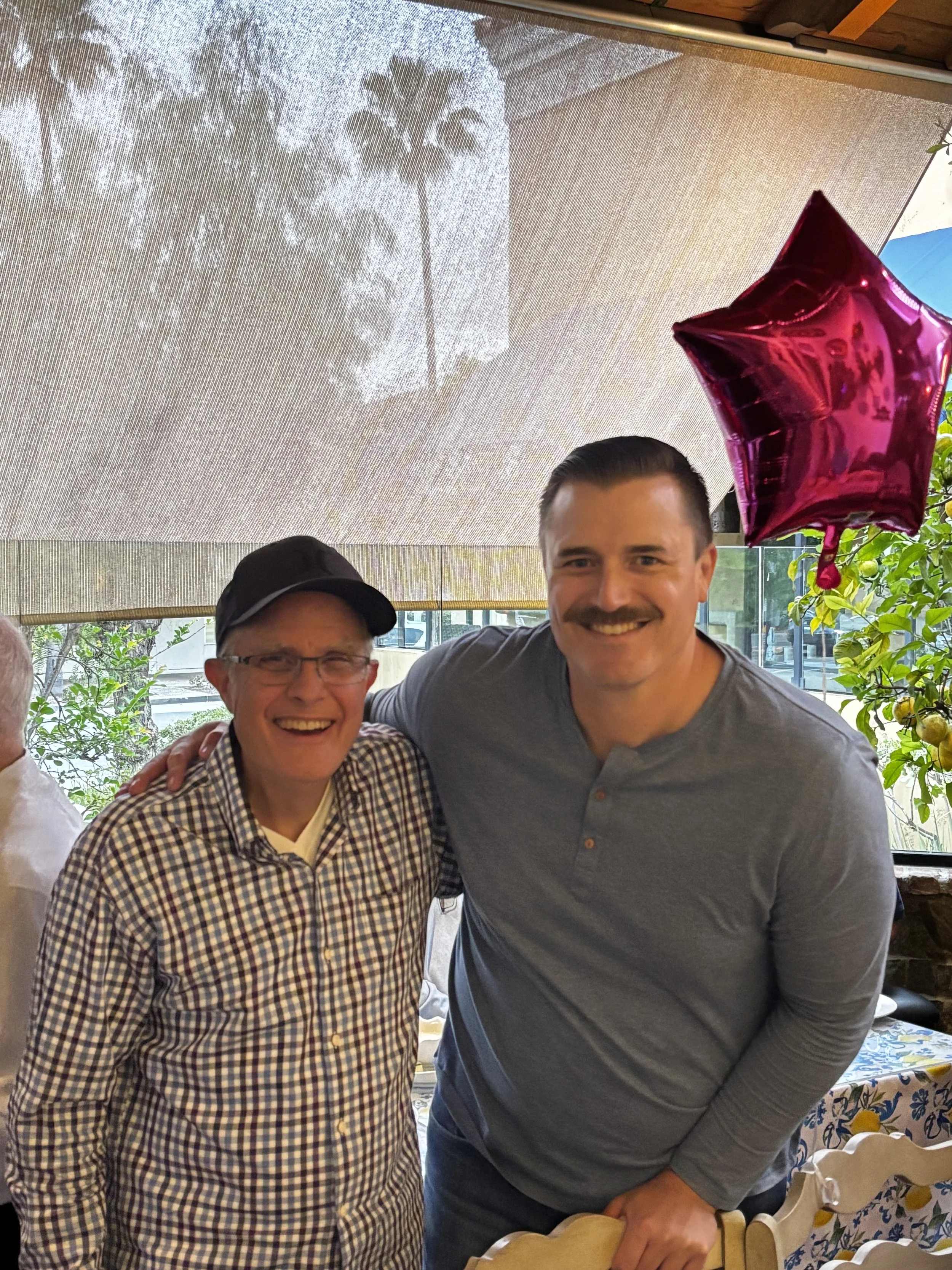Two smiling men, one older with glasses and a checkered shirt, and one younger in a gray shirt, standing close together inside a restaurant or cafe with large windows and plants in the background. A pink star-shaped balloon is visible above them.