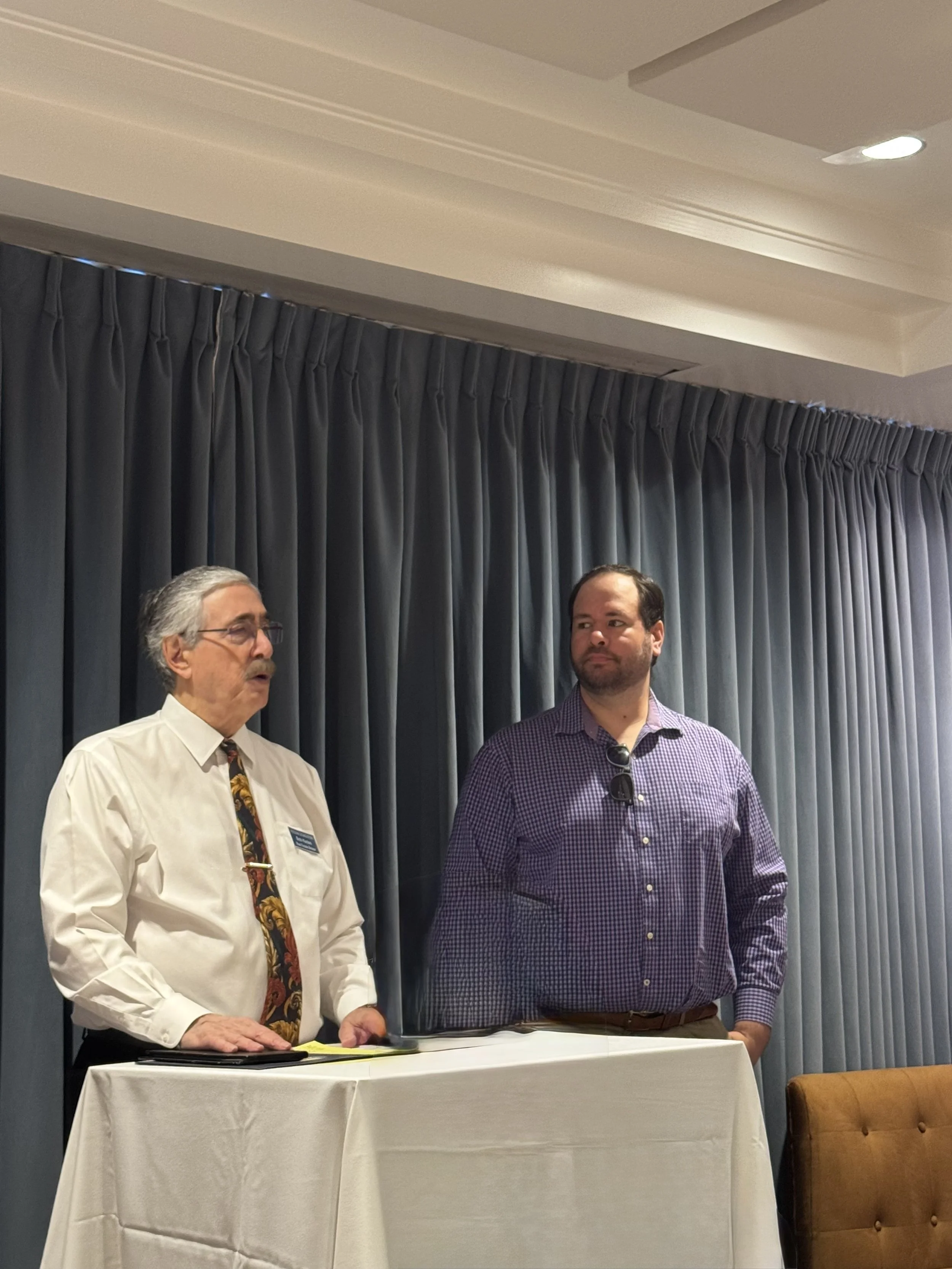 Two men are standing behind a table with a white tablecloth, appearing to participate in a formal event or discussion. The man on the left has gray hair, glasses, and is wearing a shirt with a colorful tie, while the man on the right has dark hair and is wearing a purple checkered shirt. There are dark curtains behind them, and a brown tufted chair is partially visible to the right.