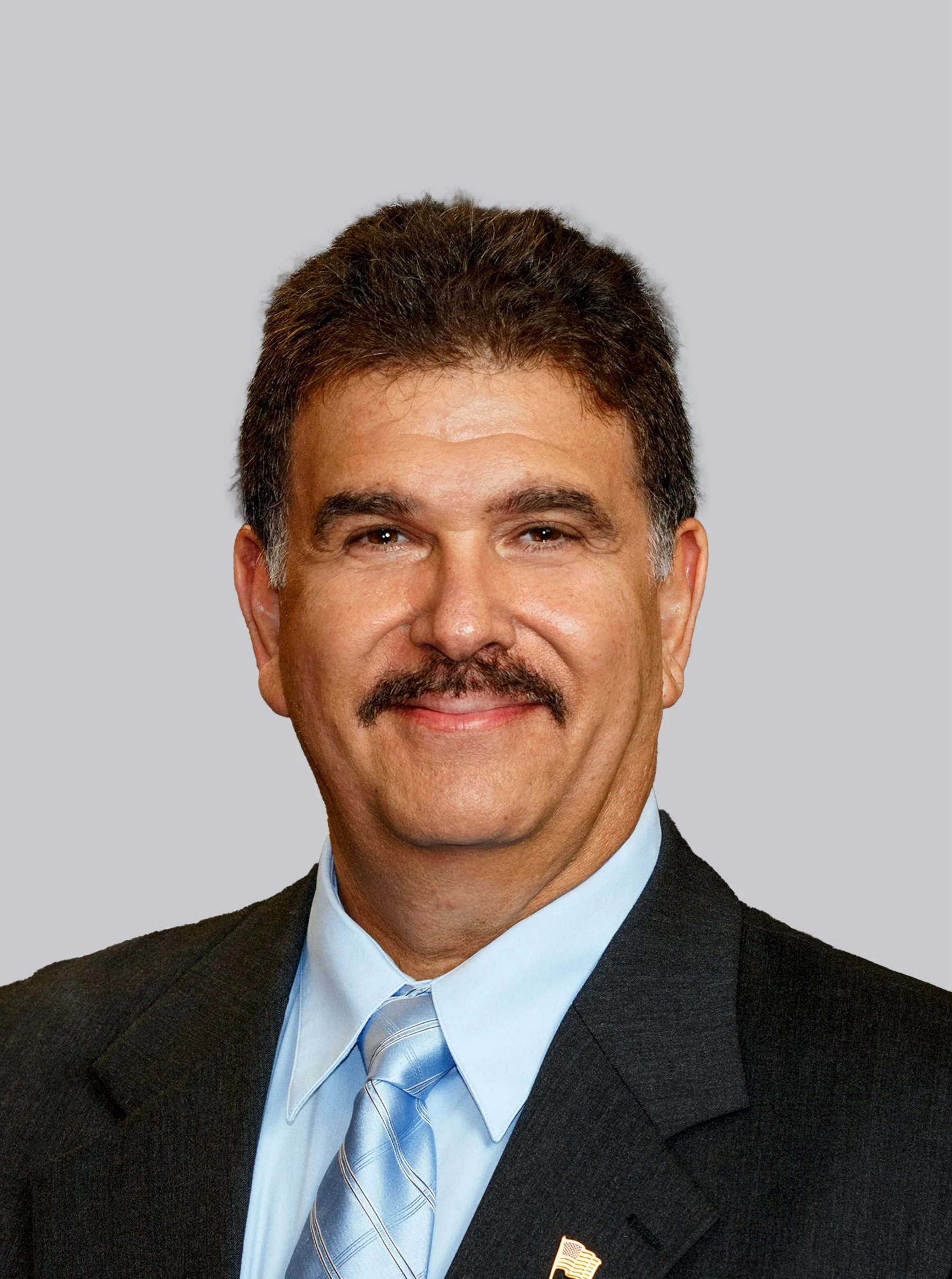 Close-up of a smiling middle-aged man with dark hair and a mustache, dressed in a suit and blue tie, wearing a pin on his jacket lapel, against a plain gray background.