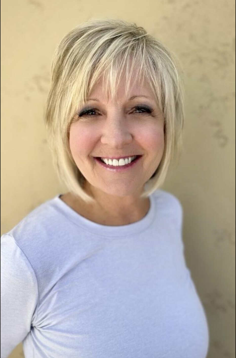 Close-up of a smiling woman with blonde hair, wearing a light gray T-shirt, standing against a beige background.