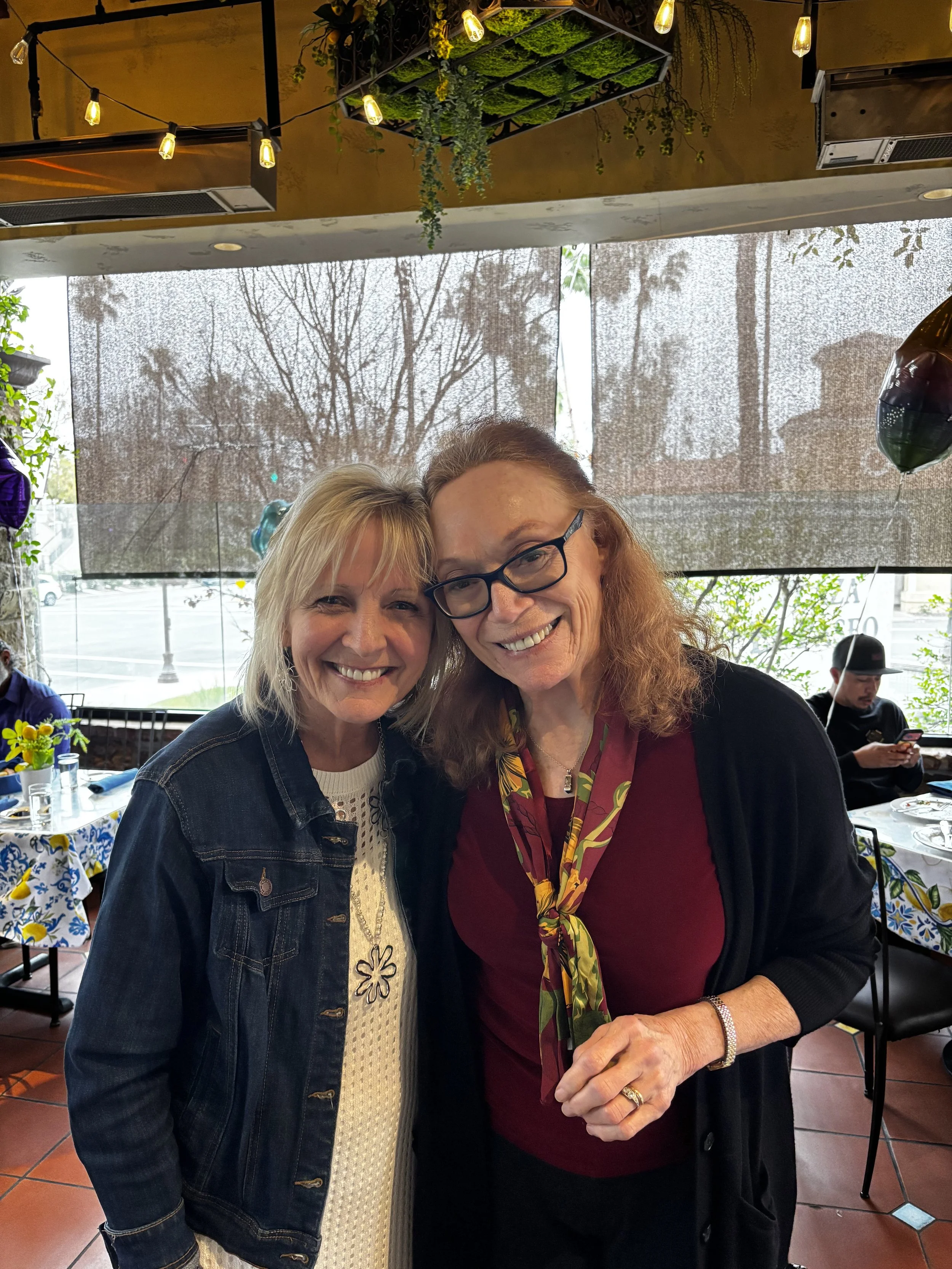 Two women are smiling and posing for a photo in a restaurant or cafe with festive decorations, including balloons and string lights, during daytime. One woman has blonde hair and is wearing a denim jacket, and the other woman has reddish hair, glasses, and a colorful scarf.