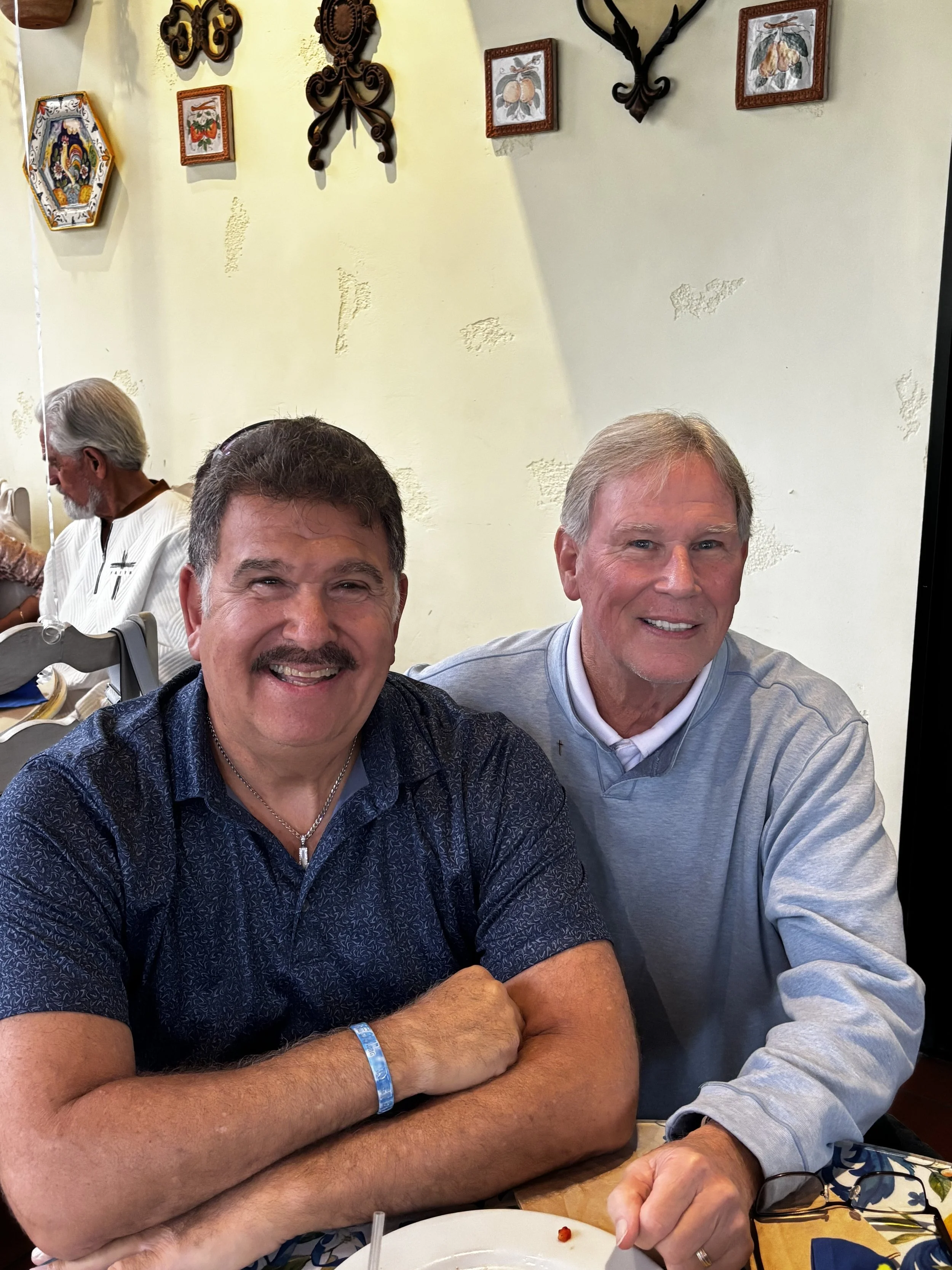 Two smiling men are sitting at a table in a restaurant, with various decorative art pieces on the yellow wall behind them. One man has a mustache and is wearing a dark patterned shirt, and the other is dressed in a light gray sweater.