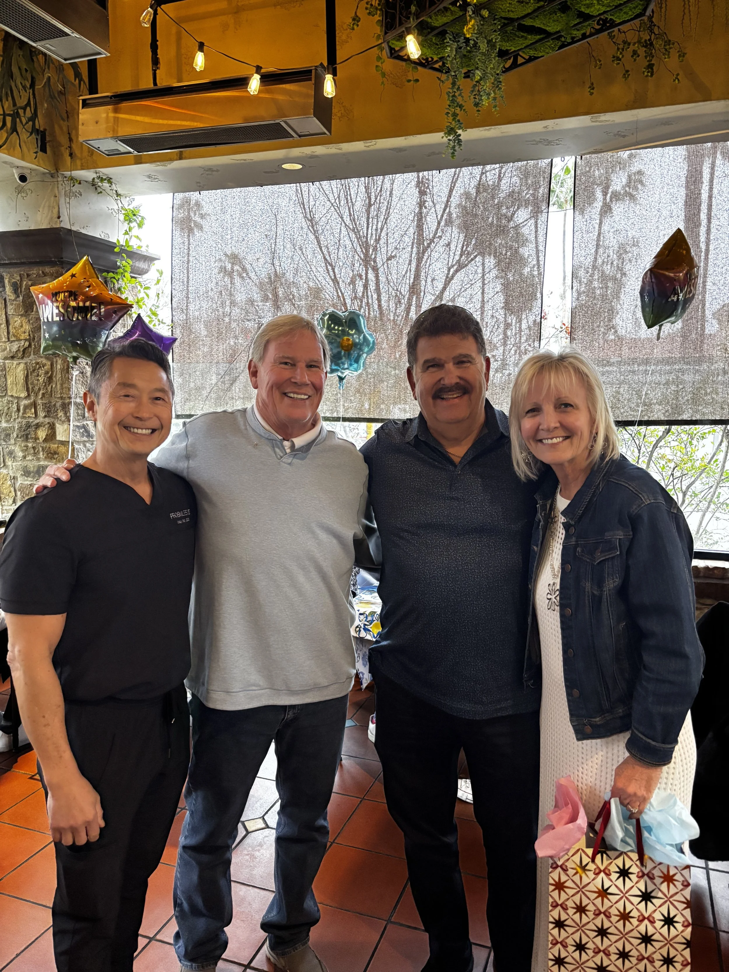 Four people standing close together, smiling in a restaurant or cafe, with balloons and gift bags in the background