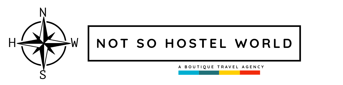 A logo for 'Not So Hostel World,' featuring a black and white compass with directions and a rectangular border around the text. Beneath the main text, there is a small line with colored segments and the phrase 'A Boutique Travel Agency.'