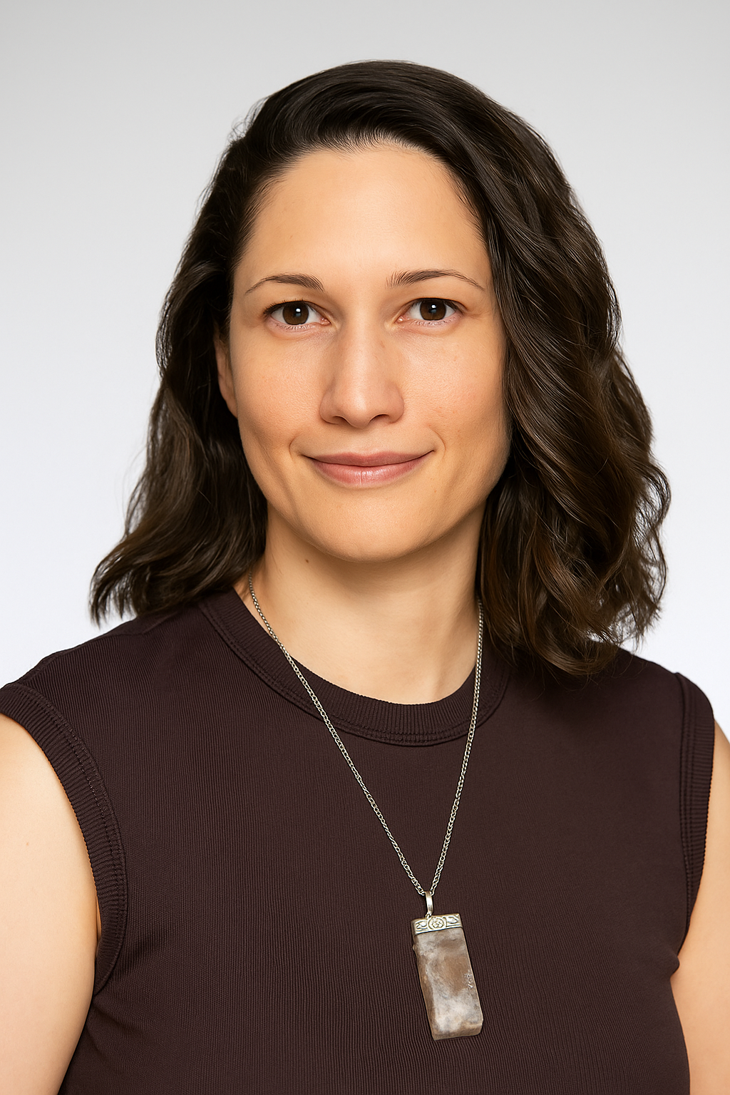 Portrait of a woman with shoulder-length brown hair, wearing a dark sleeveless top and a silver necklace with a rectangular pendant, smiling against a plain white background.