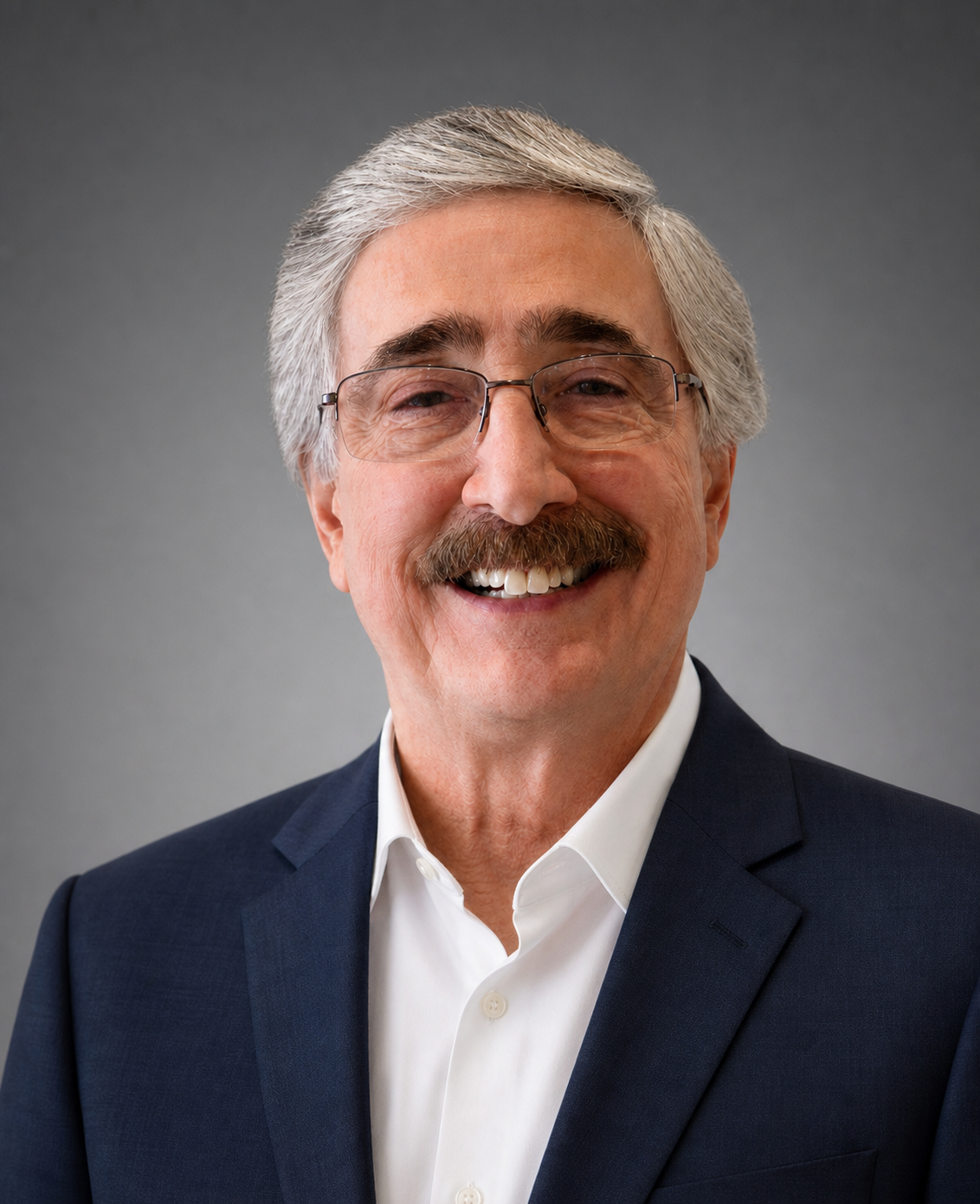 A smiling older man with gray hair, a mustache, glasses, wearing a navy blazer and white shirt, standing against a gray background.