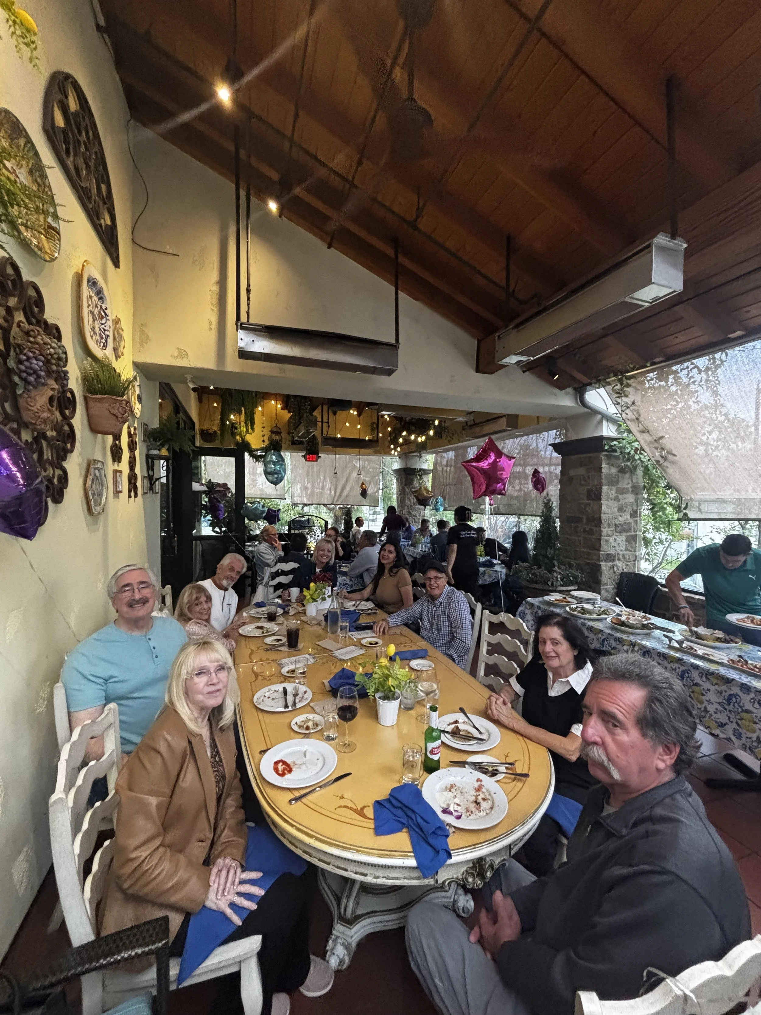 People gathered around a long dining table at a restaurant celebrating, with balloons and festive decorations, smiling and enjoying a meal.