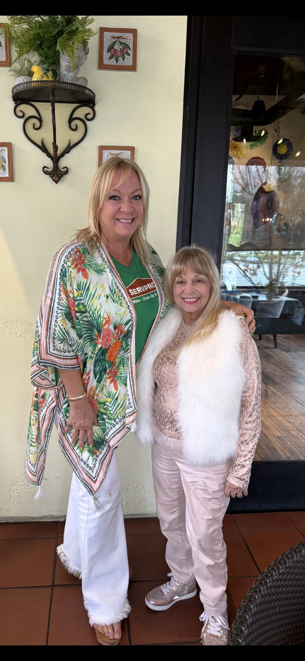 Two women smiling and posing together inside a restaurant. The taller woman on the left has blonde hair and is wearing a colorful tropical-print cover-up over a green shirt, white pants, and sandals. The shorter woman on the right has blonde hair and is wearing a beige leopard print top, a white fur vest, pink pants, and sparkly sneakers. The background includes framed botanical illustrations and a window with a view outside.