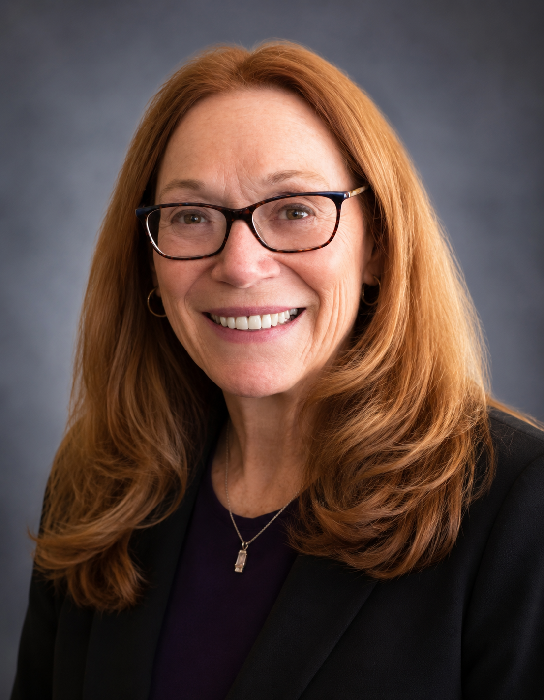 A smiling woman with long red hair, glasses, and a black blazer, wearing a necklace with a rectangular pendant, against a dark gray background.
