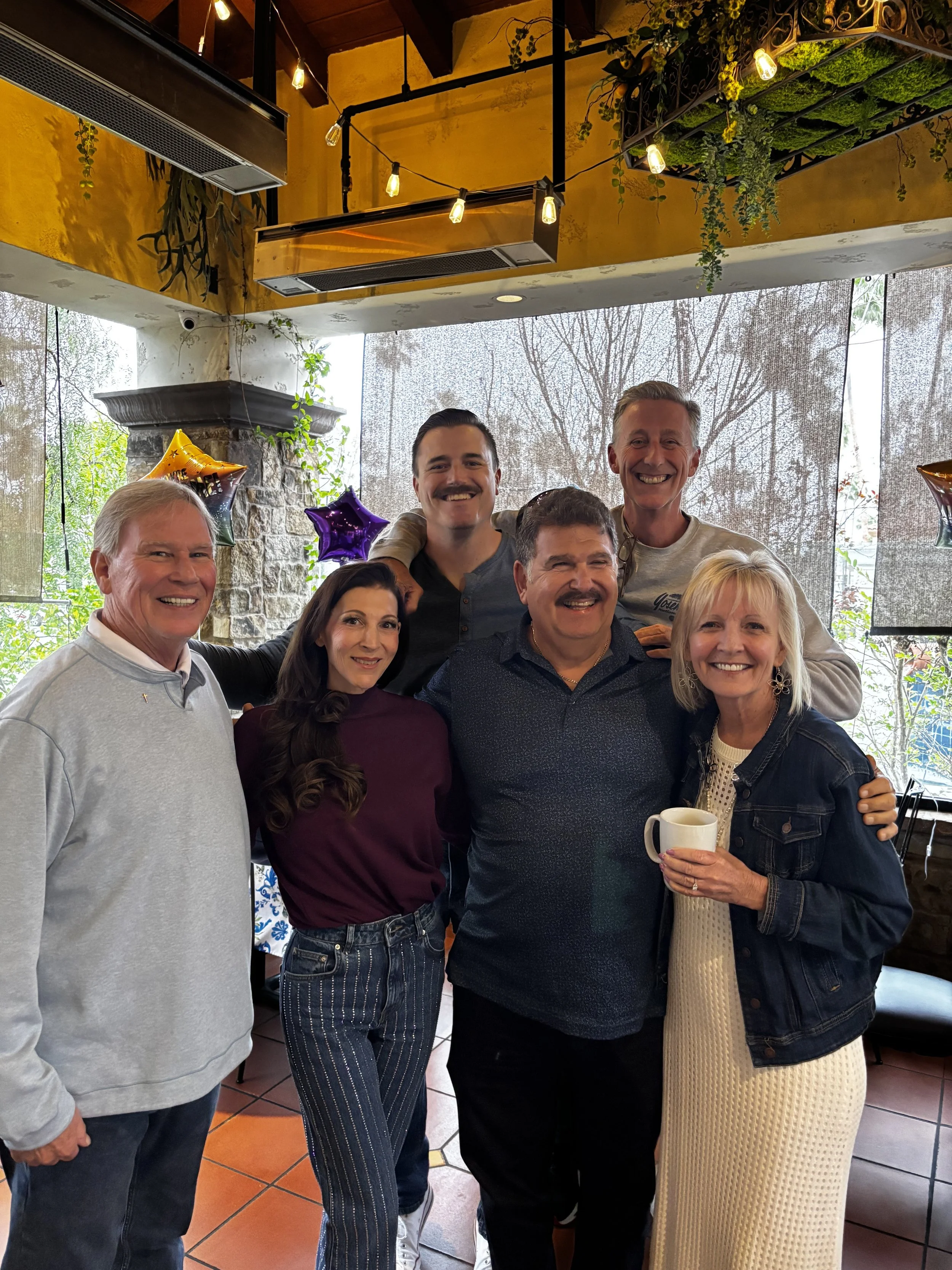 A group of seven people smiling and posing together in a restaurant or cafe with balloons and decorations, including star-shaped balloons, in the background. The setting has natural light from large windows with a scenic view outside.