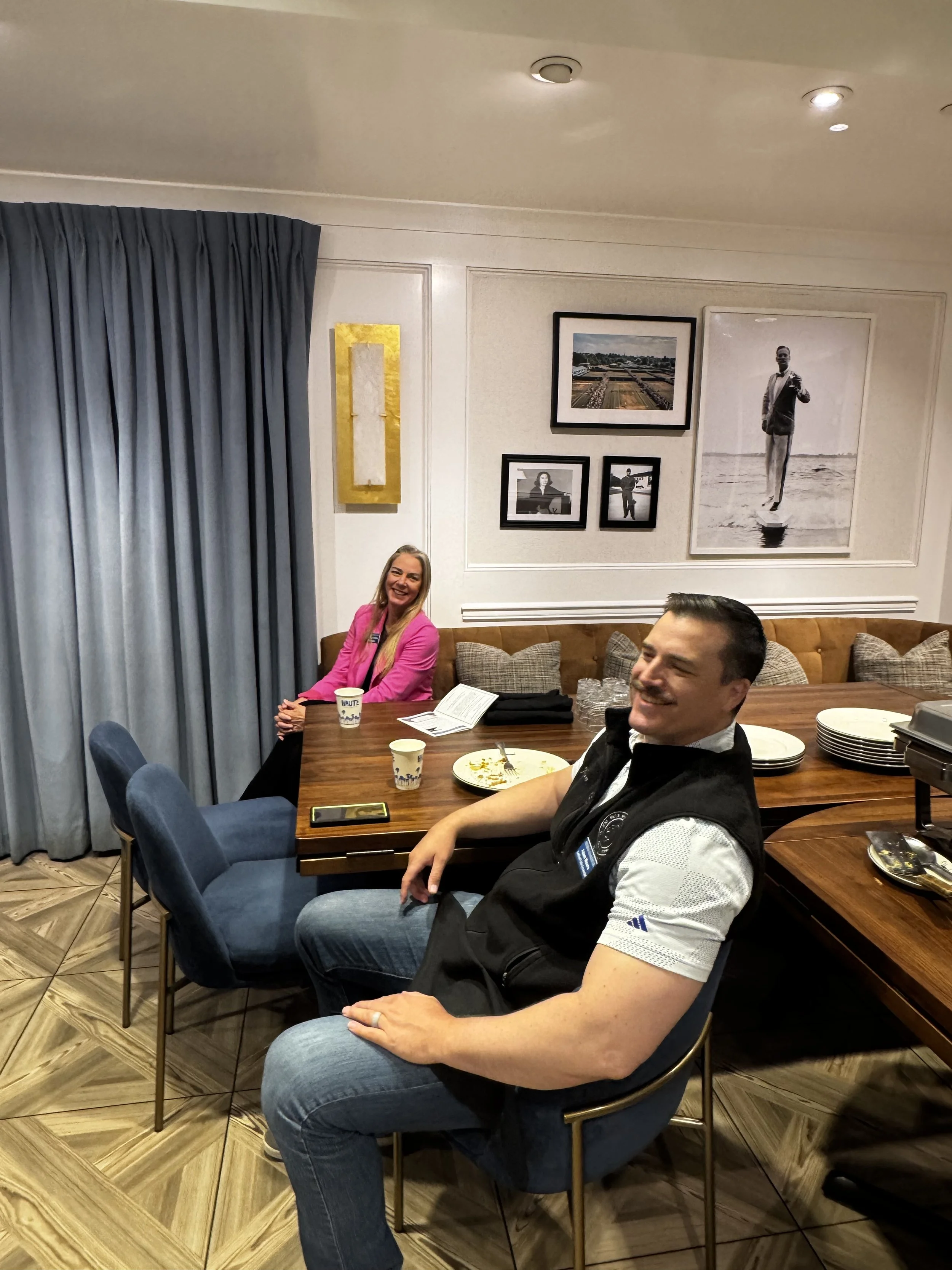 People sitting around a dining table in a restaurant or banquet hall, with framed photographs and artwork on the wall behind them. The woman is wearing a bright pink blazer and smiling, while the man is wearing a black vest over a white shirt and also smiling.