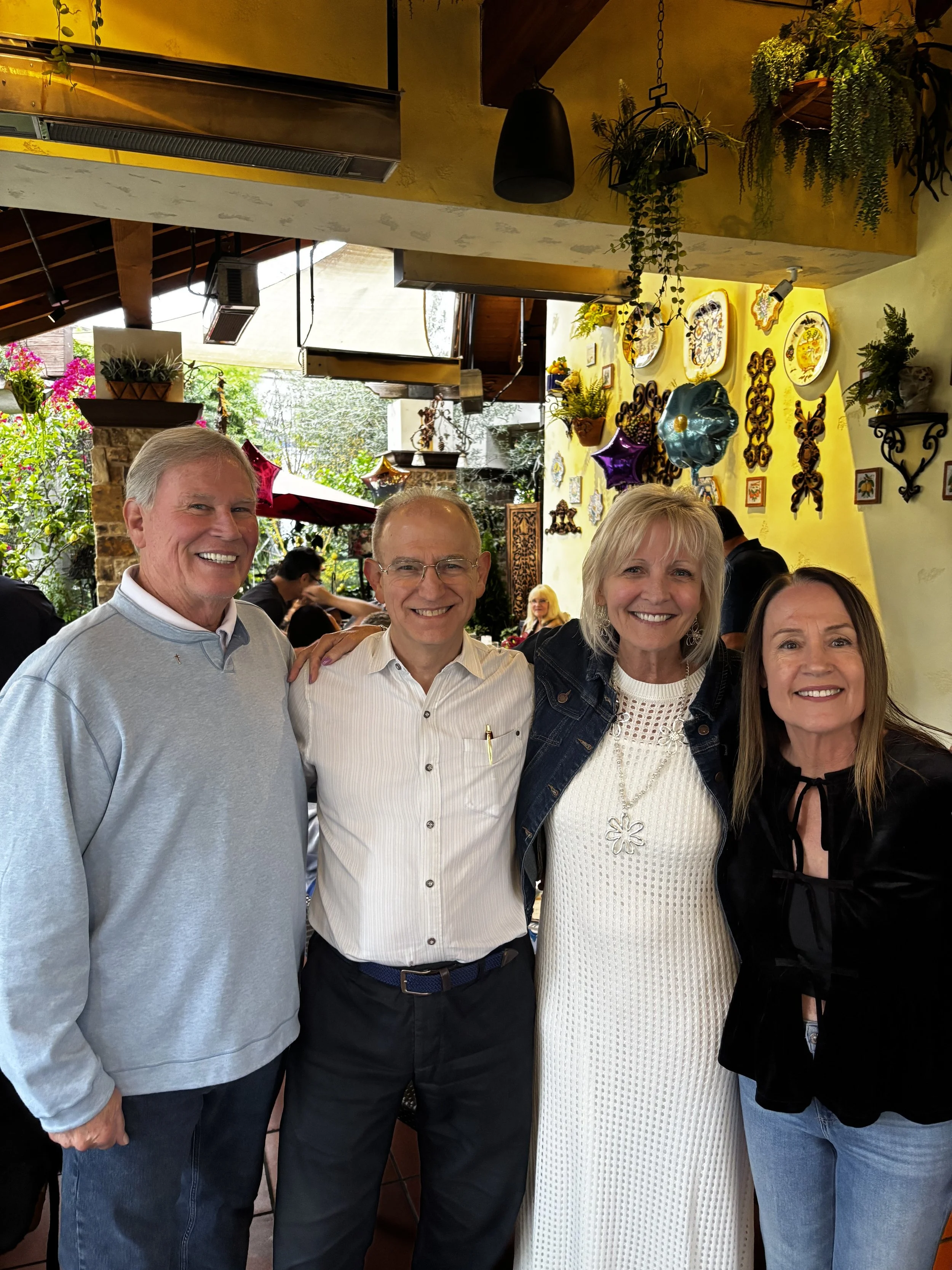 Four smiling adults, two men and two women, standing close together in an indoor setting decorated with hanging plants, colorful plates, and balloons.