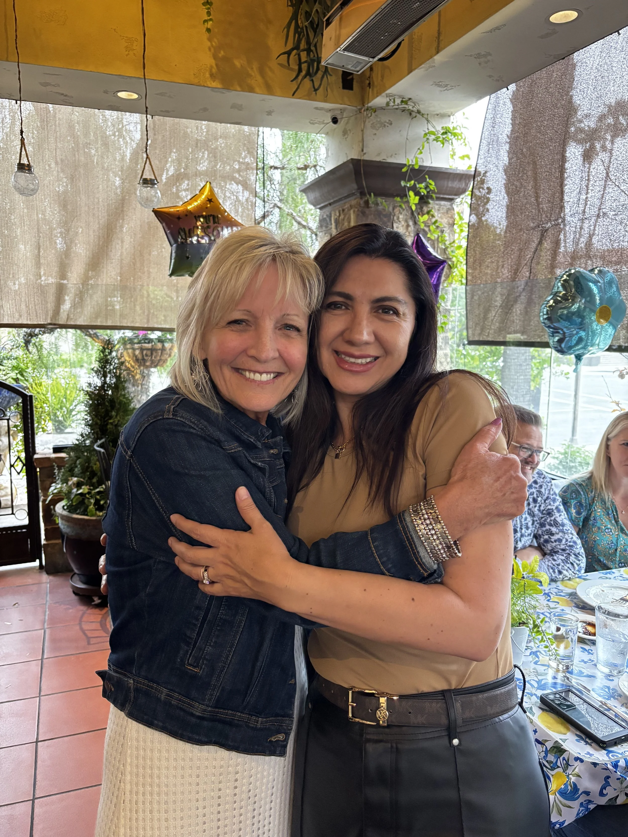 Two women hugging and smiling at a celebration or gathering in a restaurant or cafe, with balloons and other people in the background.