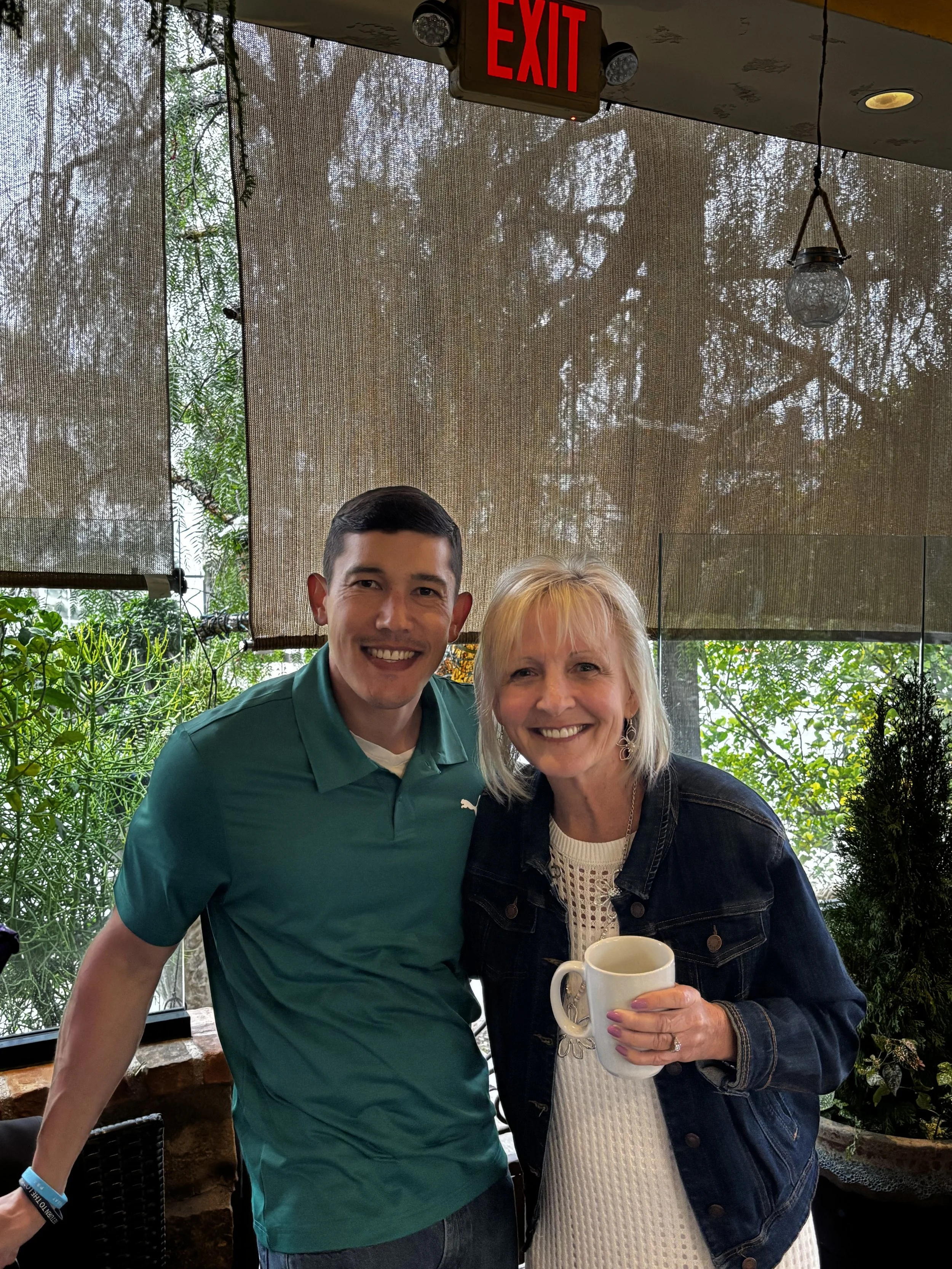 A young man and an older woman are smiling and posing together indoors. The woman is holding a white mug. The background shows trees and greenery outside a large window, with a red EXIT sign at the top.