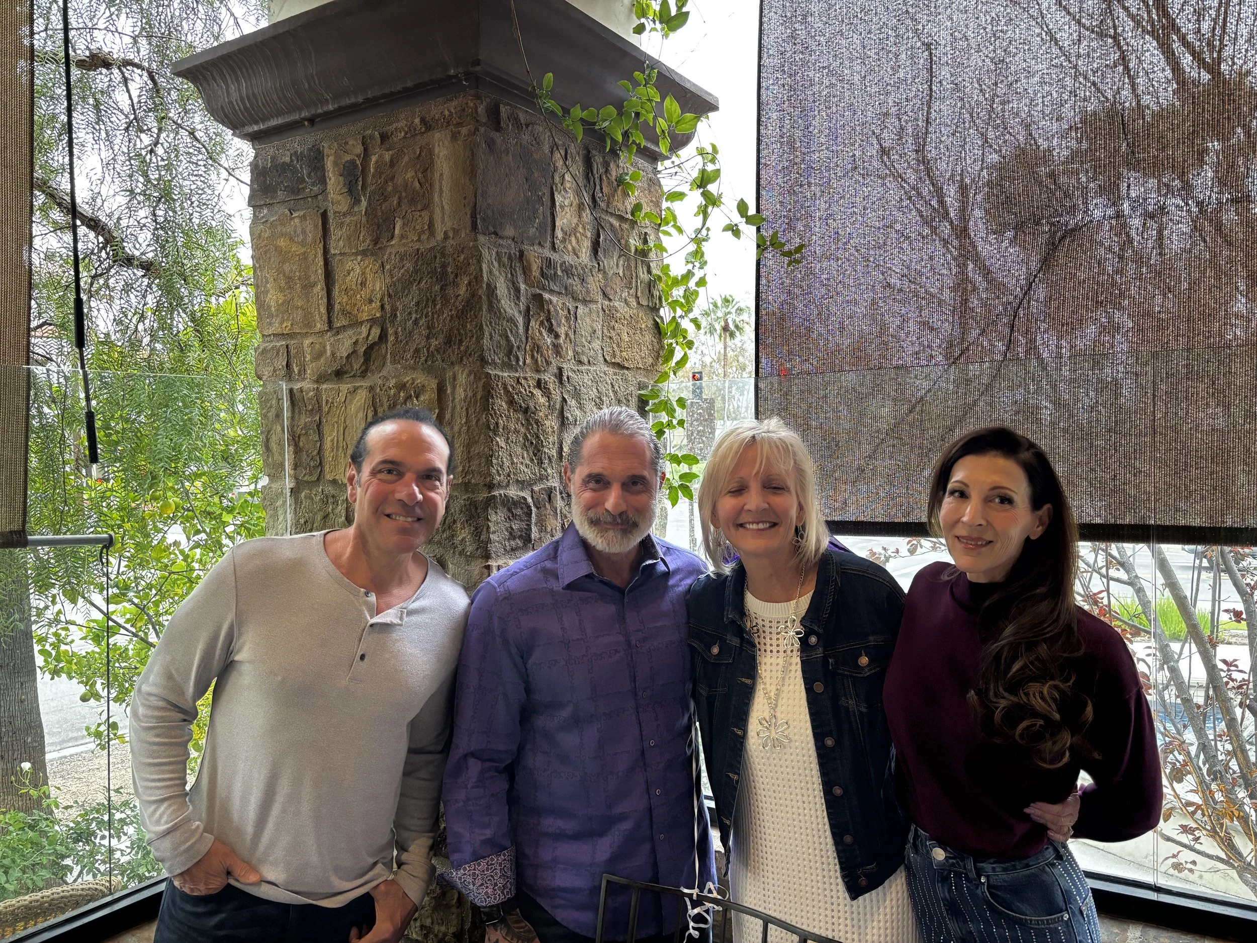 Four people standing closely together indoors, with trees outside and a stone pillar behind them.