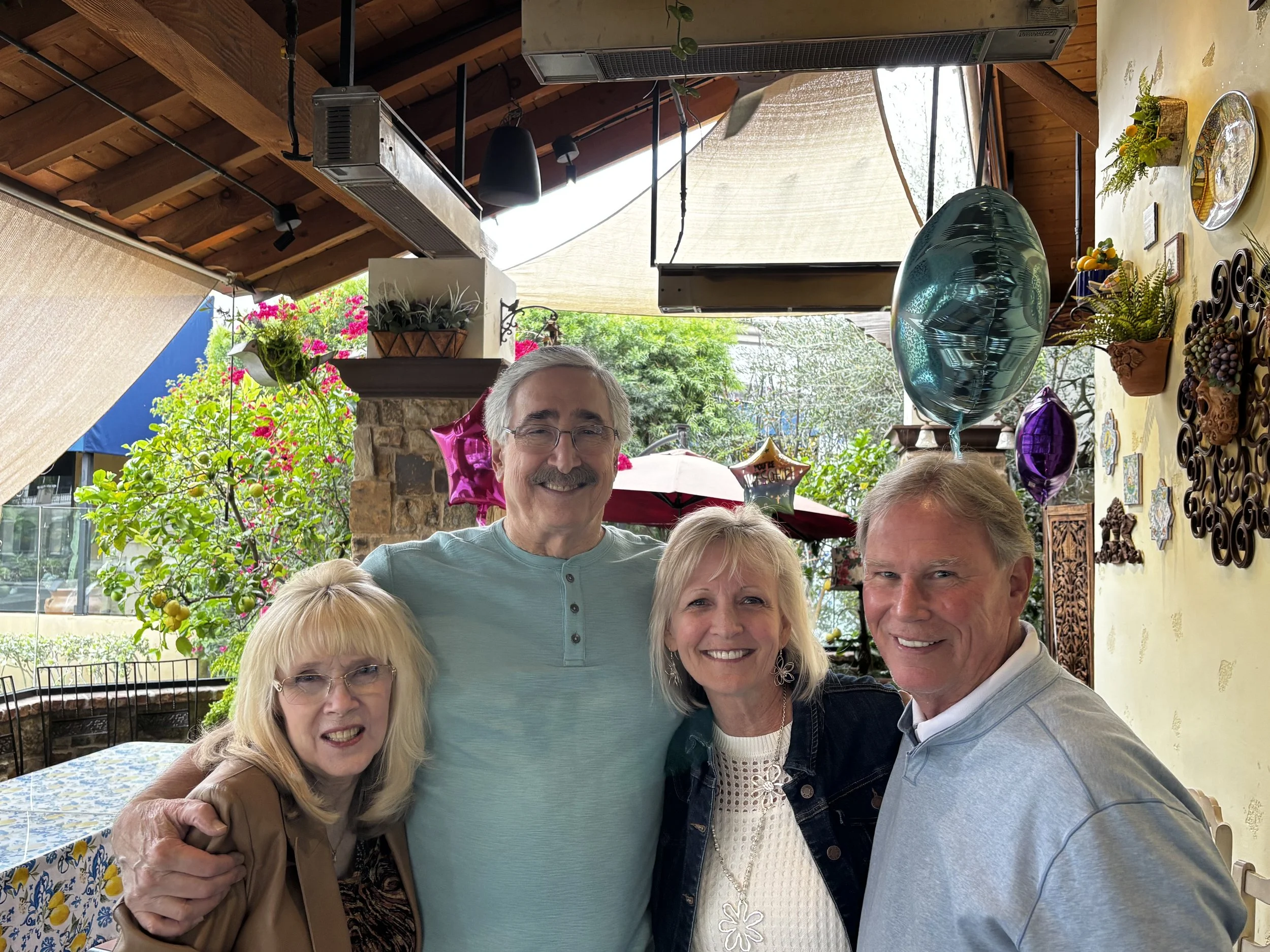 Group of five smiling people gathered outdoors under a covered patio, surrounded by colorful balloons and decorative items, with a lush garden and flowering trees in the background.