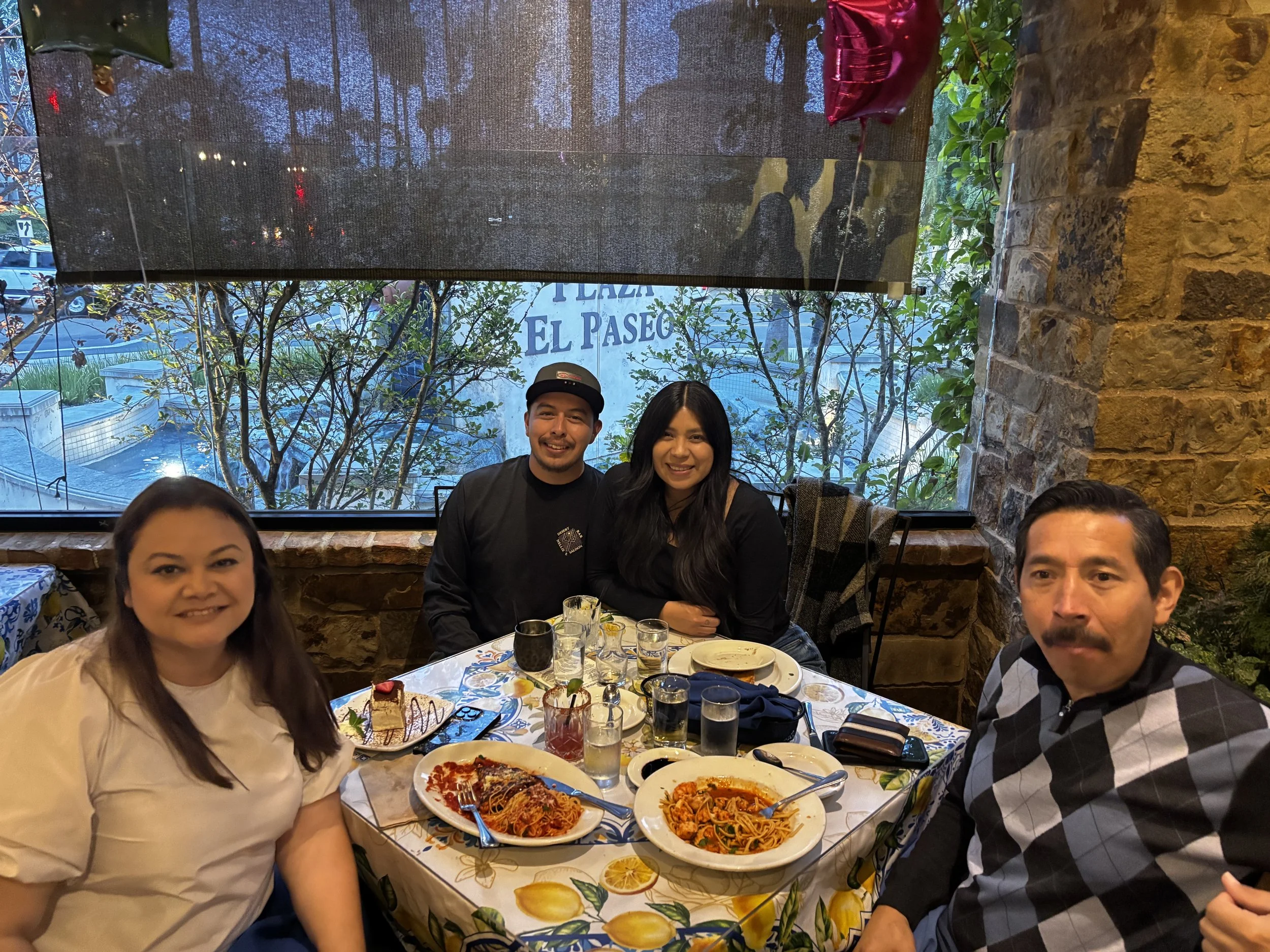 A group of people sitting around a table in a restaurant, enjoying Italian food such as pizza and pasta, with a large window showing trees and outdoor scenery in the background.