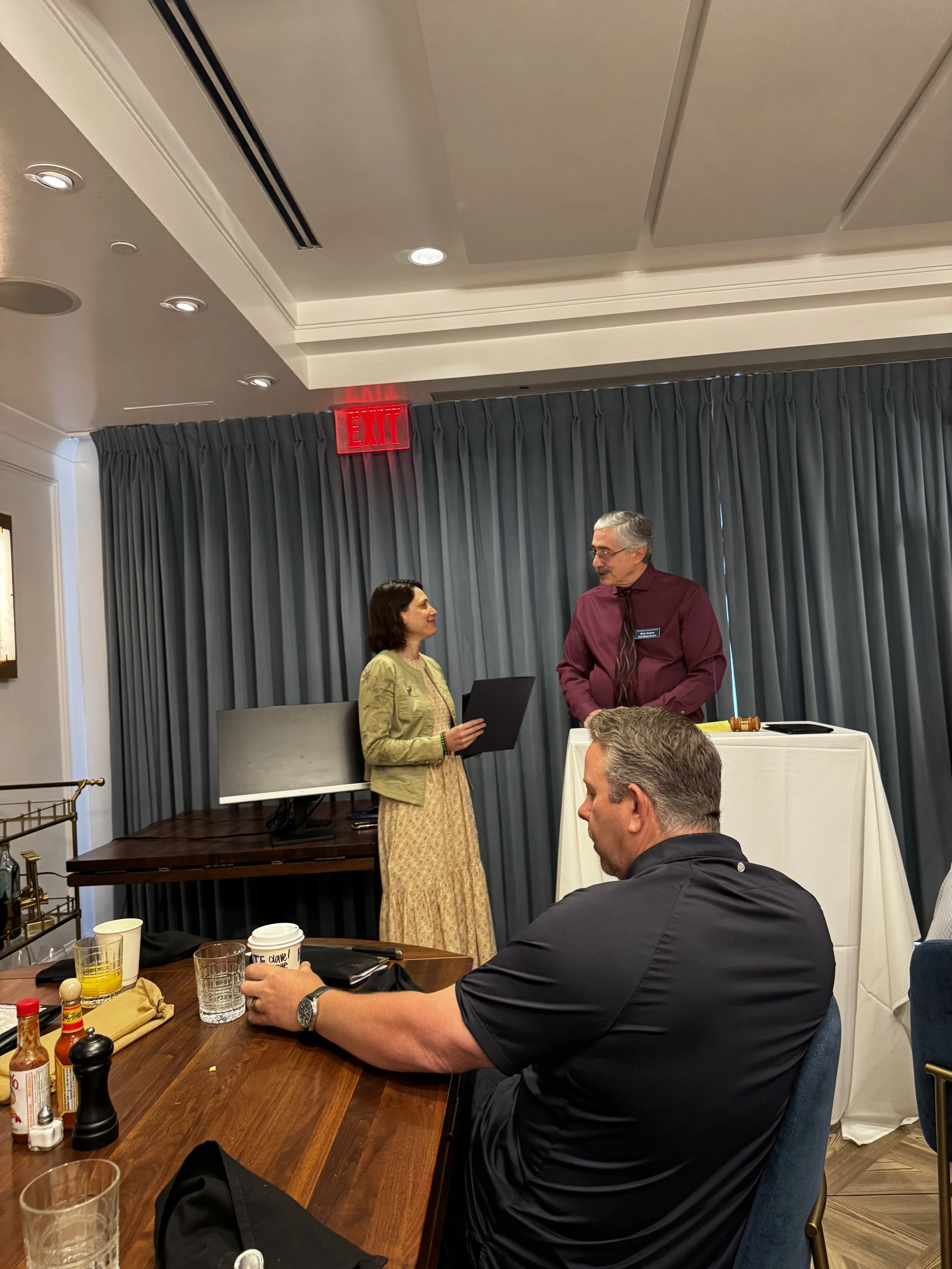 Two people stand in front of a curtain, engaged in conversation at an indoor event; a table with glasses and condiments is visible in the foreground, and a man with gray hair, glasses, and a maroon shirt is speaking to a woman in a beige dress holding a folder.