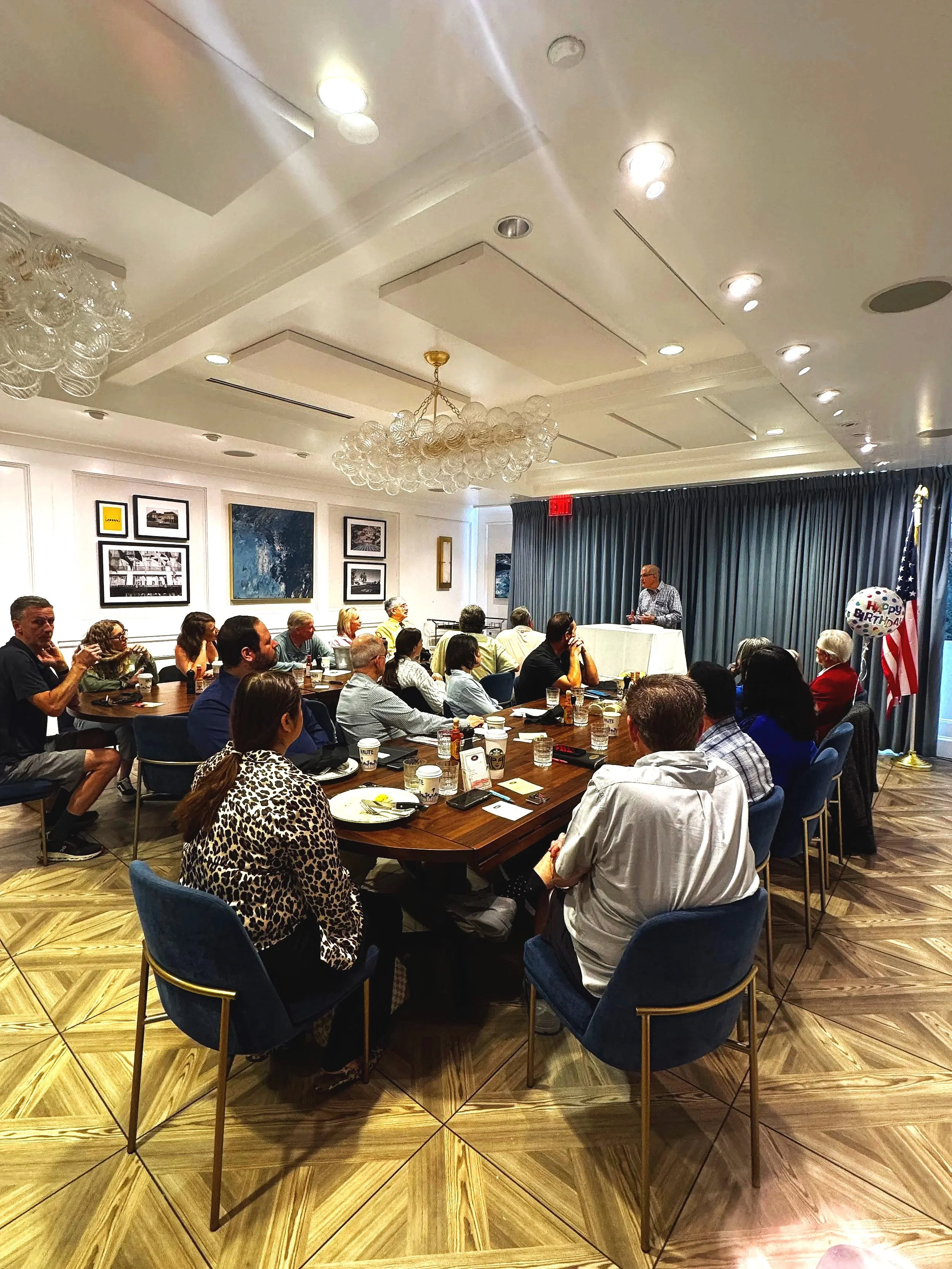 A group of diverse people attending a formal event or conference in a room with dark curtains, framed pictures on the wall, and a chandelier. A man is speaking at a podium, while others sit at tables with beverages and snacks, listening attentively.