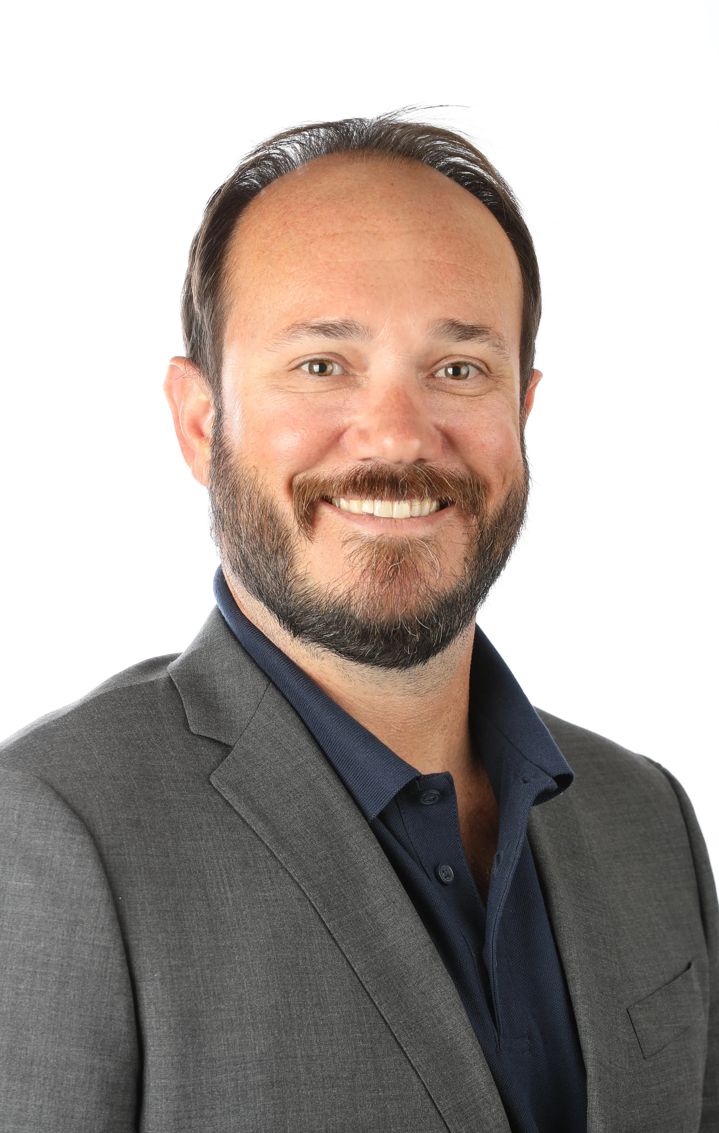 A smiling man with a beard and mustache wearing a gray suit and a dark blue shirt, looking directly at the camera against a plain white background.