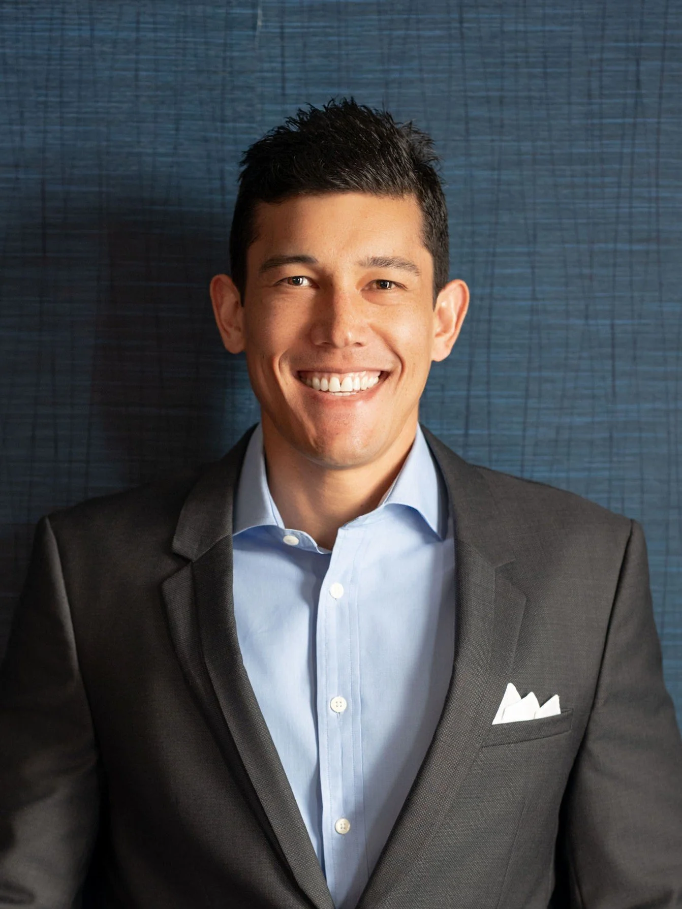 Headshot of a smiling man in a suit and light blue shirt, standing against a blue textured background.