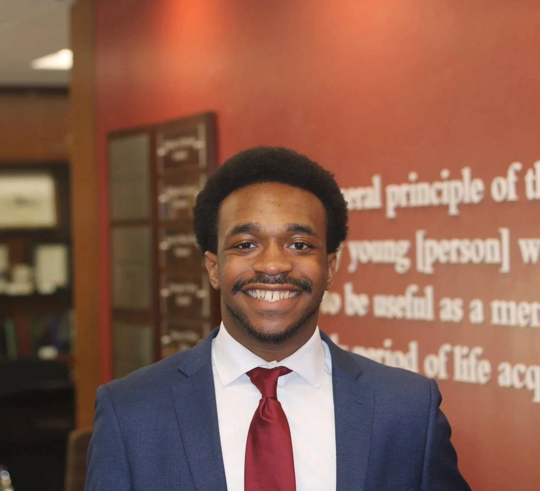 Young man dressed in a navy suit, white shirt, and red tie standing inside a building with a red wall and framed pictures or posters in the background, smiling at the camera.