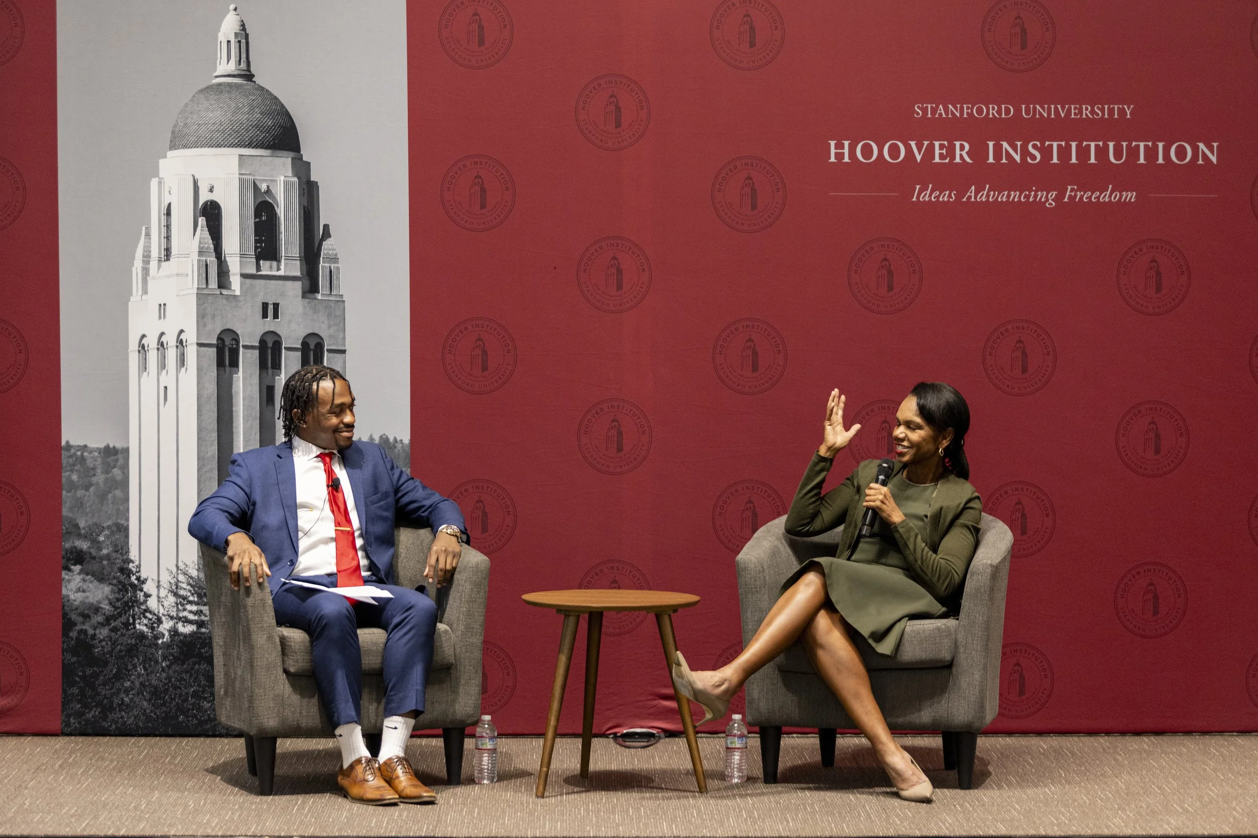 A man in a blue suit and red tie, and a woman in a green dress, are sitting on chairs on stage, engaged in a discussion or interview at Stanford University, with a red backdrop and Harvard Institution branding. The woman is gesturing with her hand, and the man is smiling. There are water bottles on the floor in front of them.