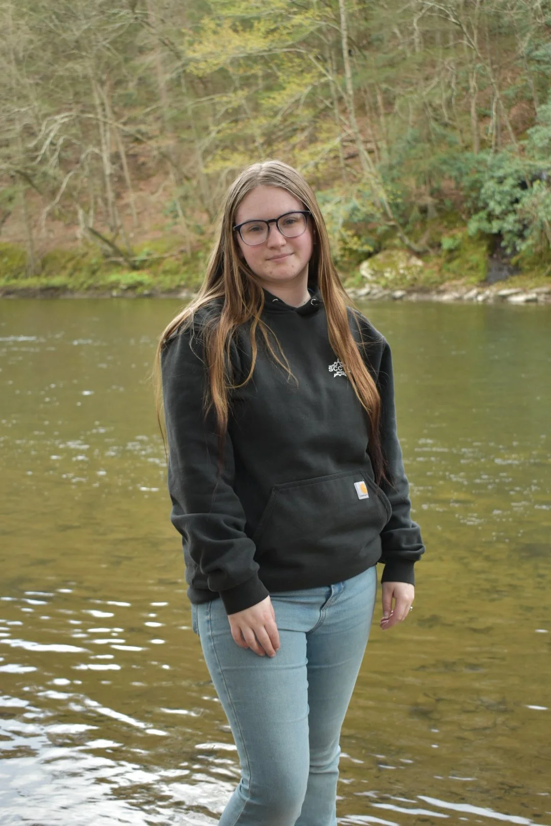 A young woman with long brown hair and glasses standing in a river with a wooded area in the background.