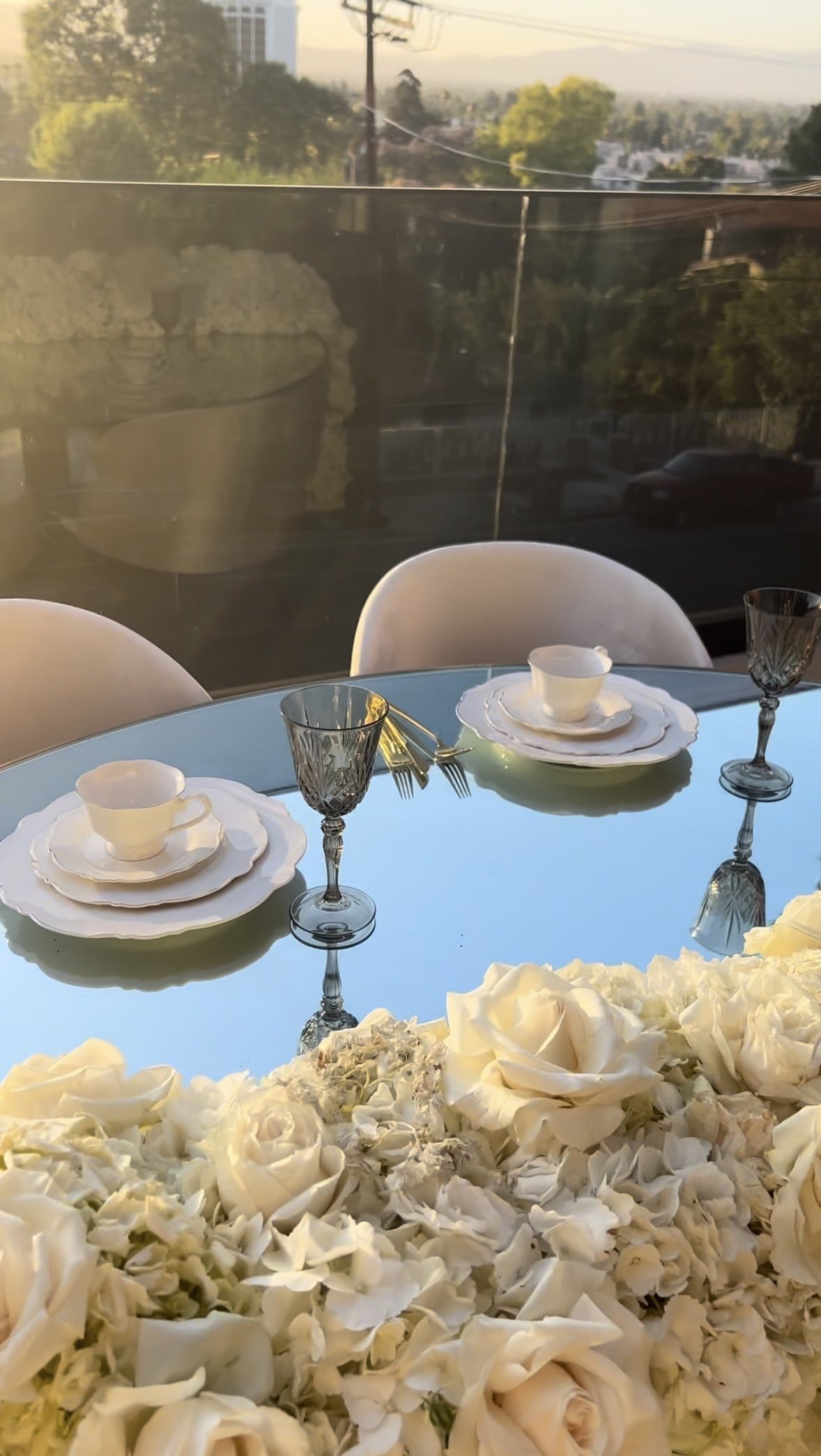 Elegant table setting with white plates, teacups, and crystal wine glasses on a mint green table, with a background view of a cityscape and trees, and a large flower arrangement of white roses and petals in the foreground.