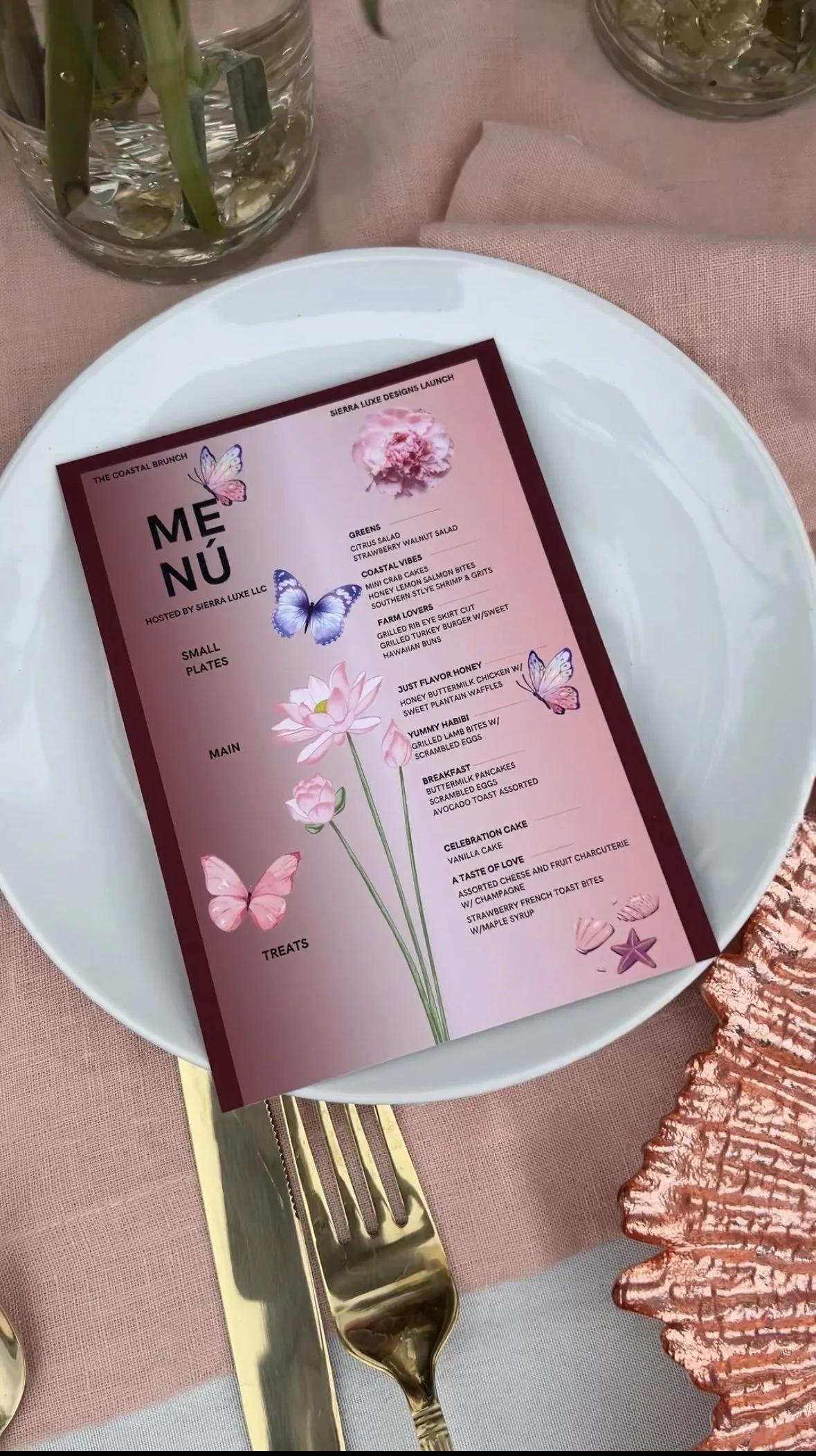 A pink menu with floral and butterfly designs on a white plate, with gold utensils and a pink napkin on a table with pink tablecloths and glass vases in the background.