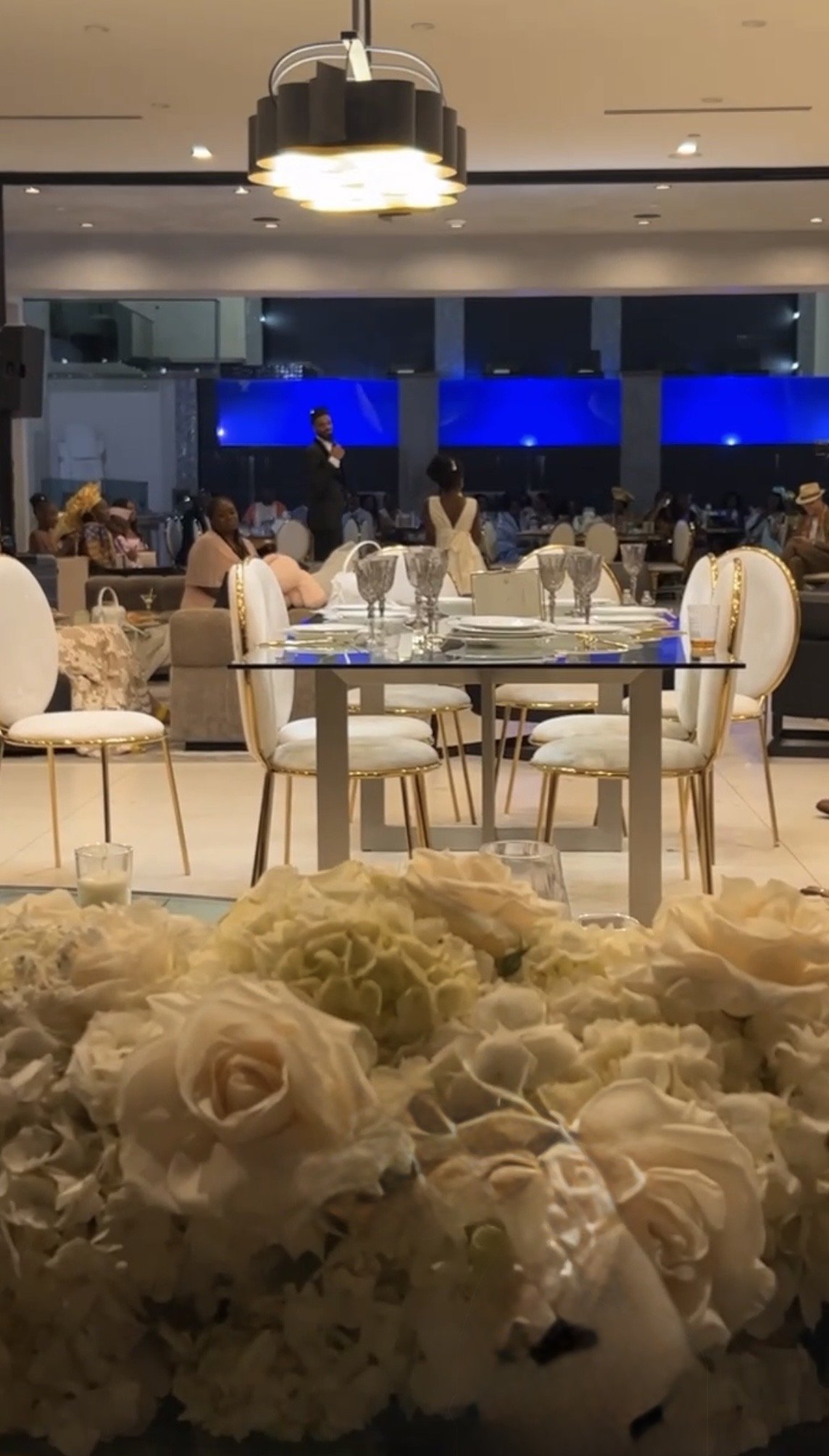 Indoor banquet hall with elegant white chairs, a table set with plates and glasses, and a floral centerpiece at the front. In the background, a man and woman are standing and addressing the guests. The room has large blue screens and warm lighting.