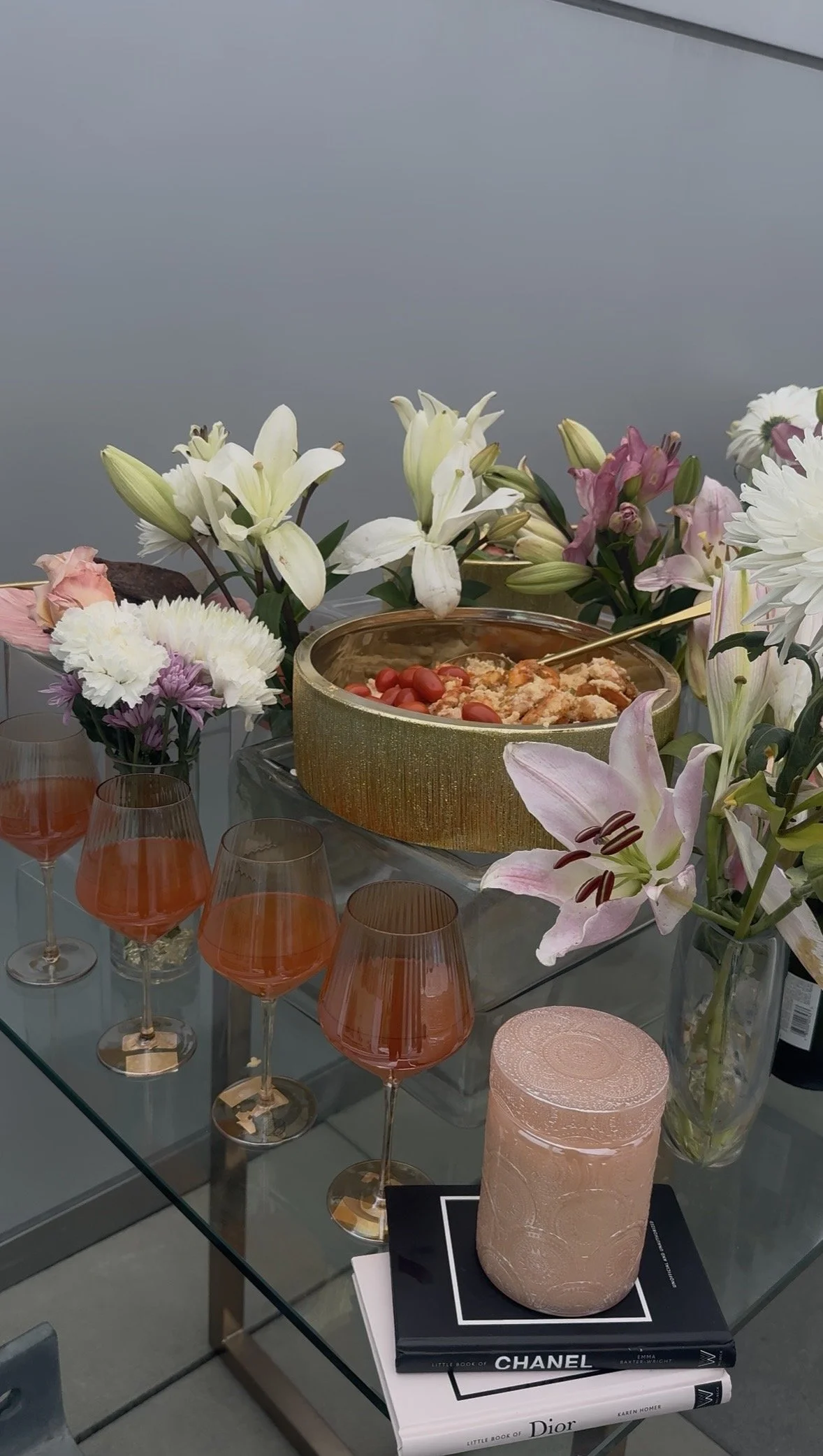 Table decorated with white, pink, and purple flowers, a large bowl of salad with cherry tomatoes, glasses filled with pink drink, a pink candle, and fashion books including Chanel and Dior.