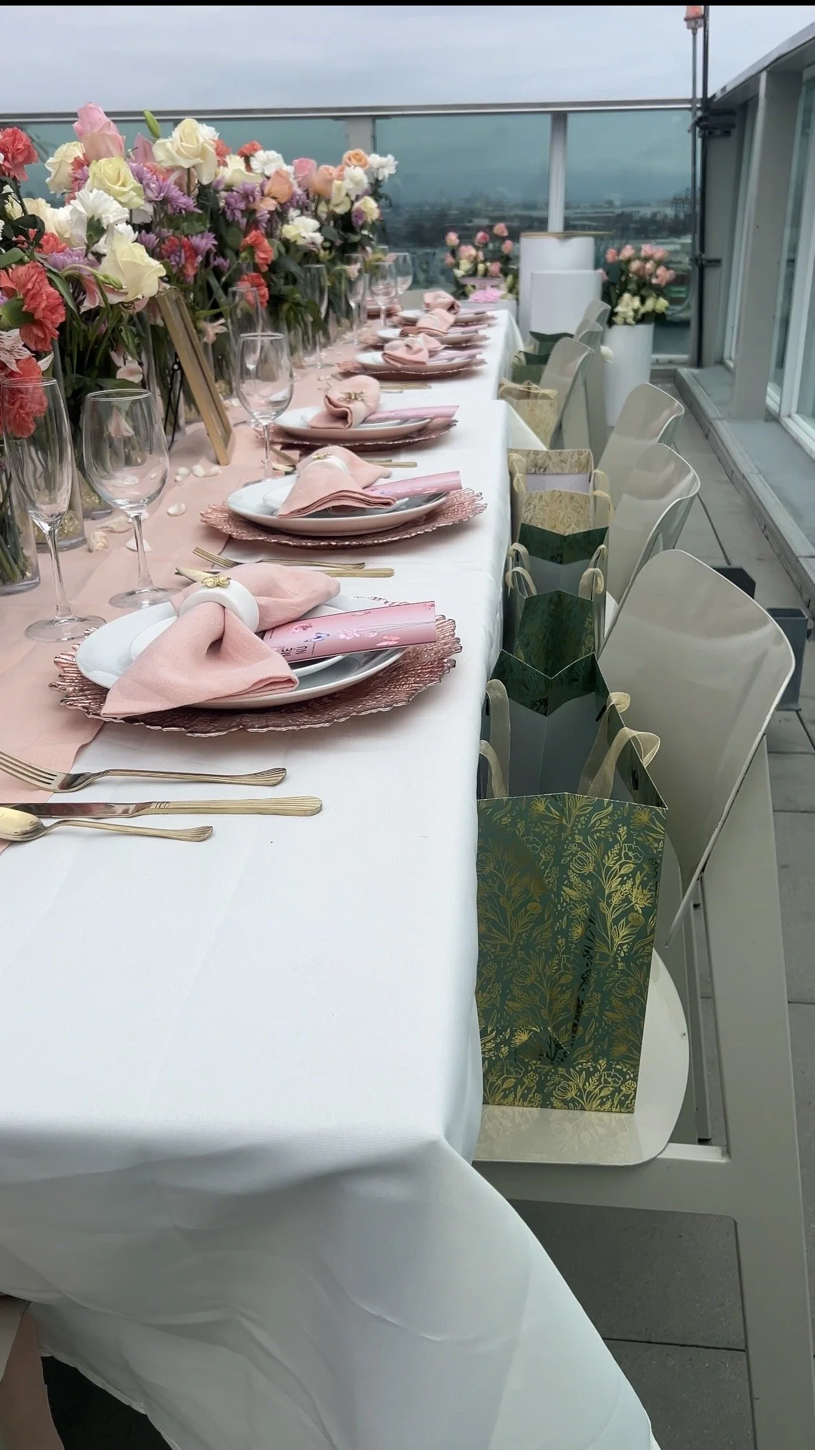 A long table decorated with pink and white flowers, set for a celebration or wedding with plates, pink napkins, glasses, and gift bags along the side, on a balcony with a view of a cityscape in the background.