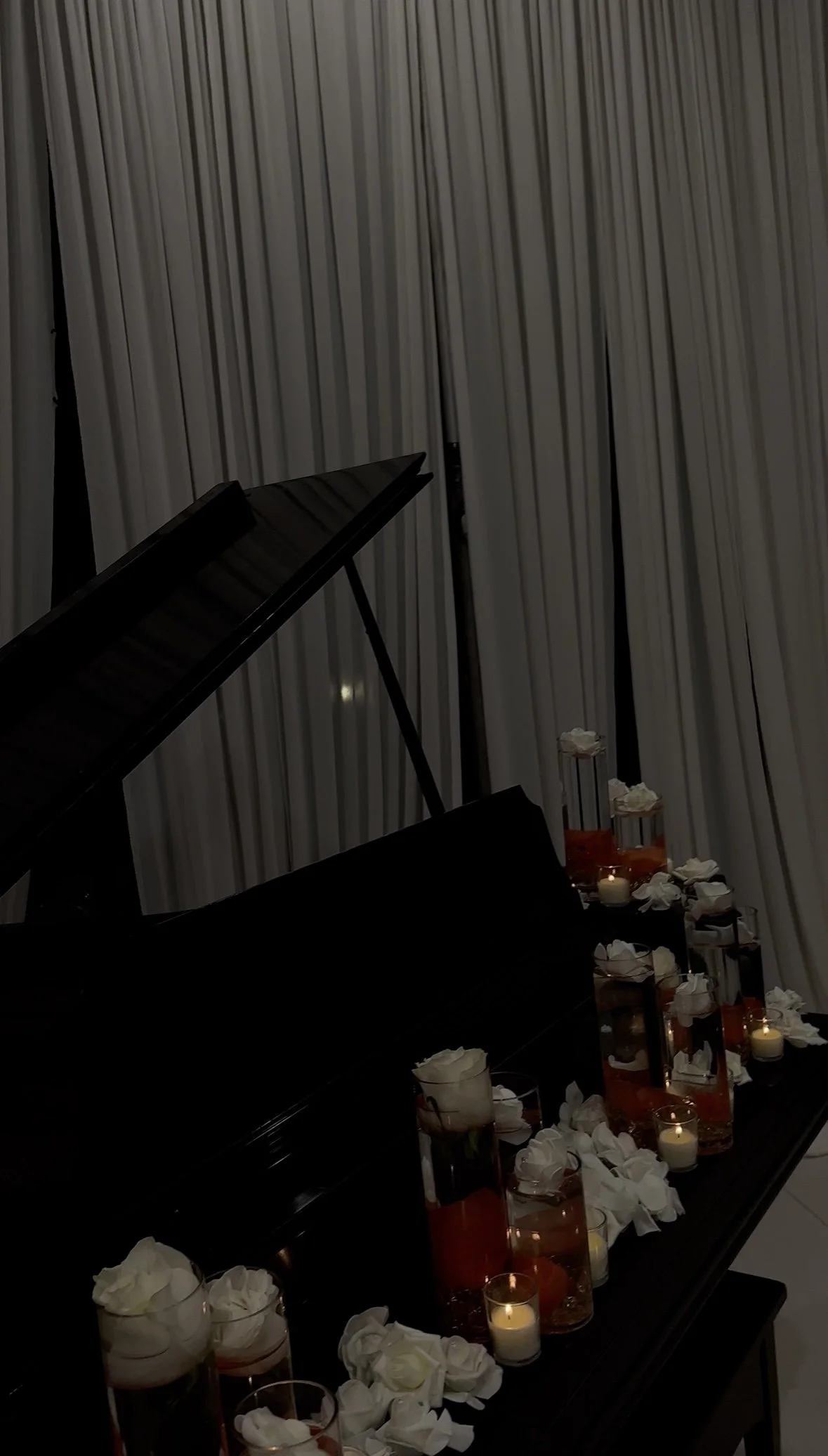 A black grand piano with a closed lid decorated with white roses and small candles in glass containers, set against a backdrop of floor-length light-colored curtains.