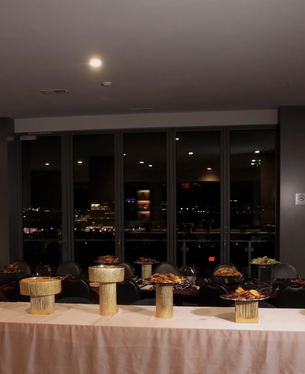 Table with multiple plates of food, likely at a catered event, with a city skyline visible through large glass windows at night.